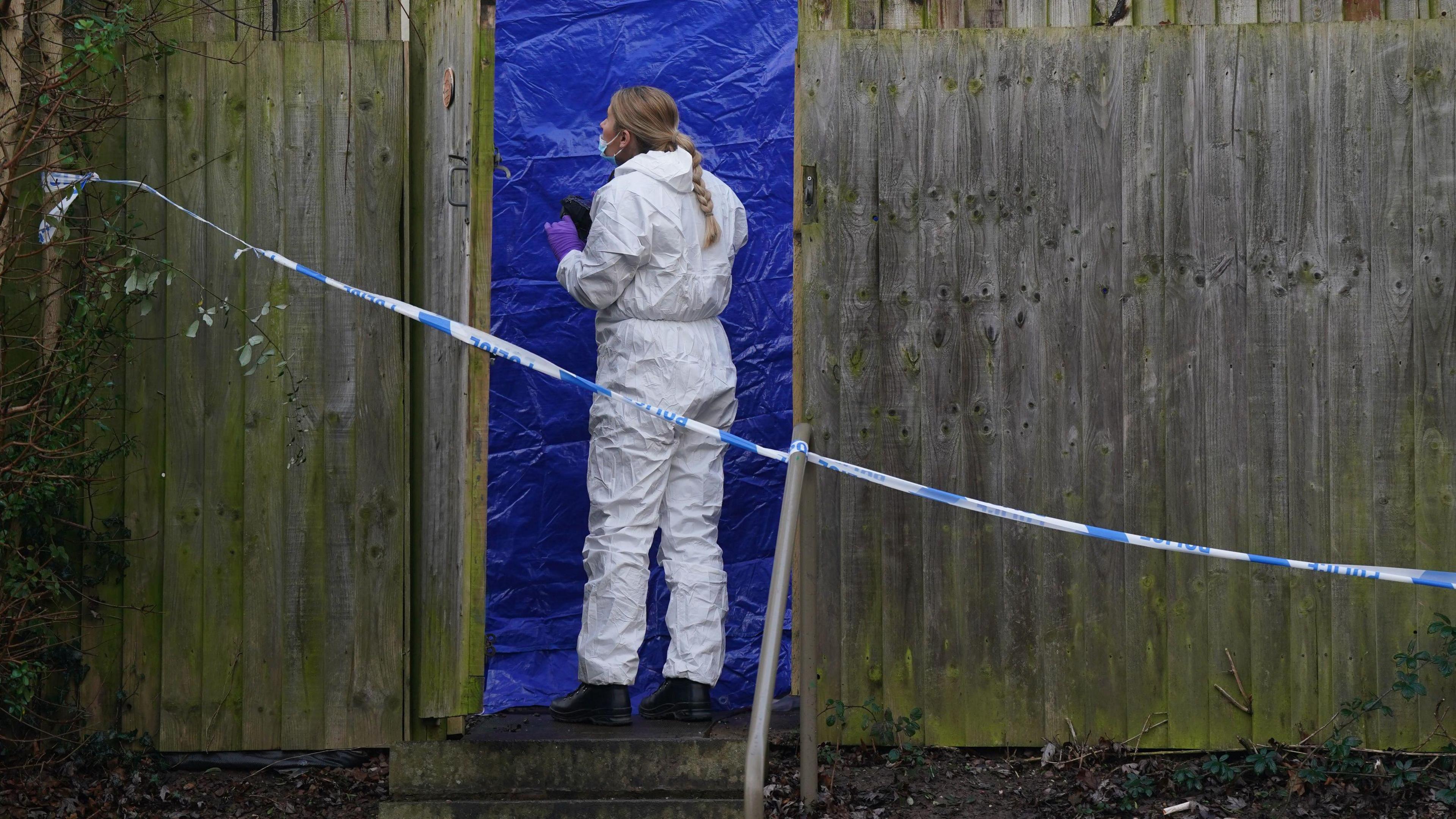 A forensic officer in a white suit standing next to a blue tent. She is behind police tape in a garden obscured by a fence. 