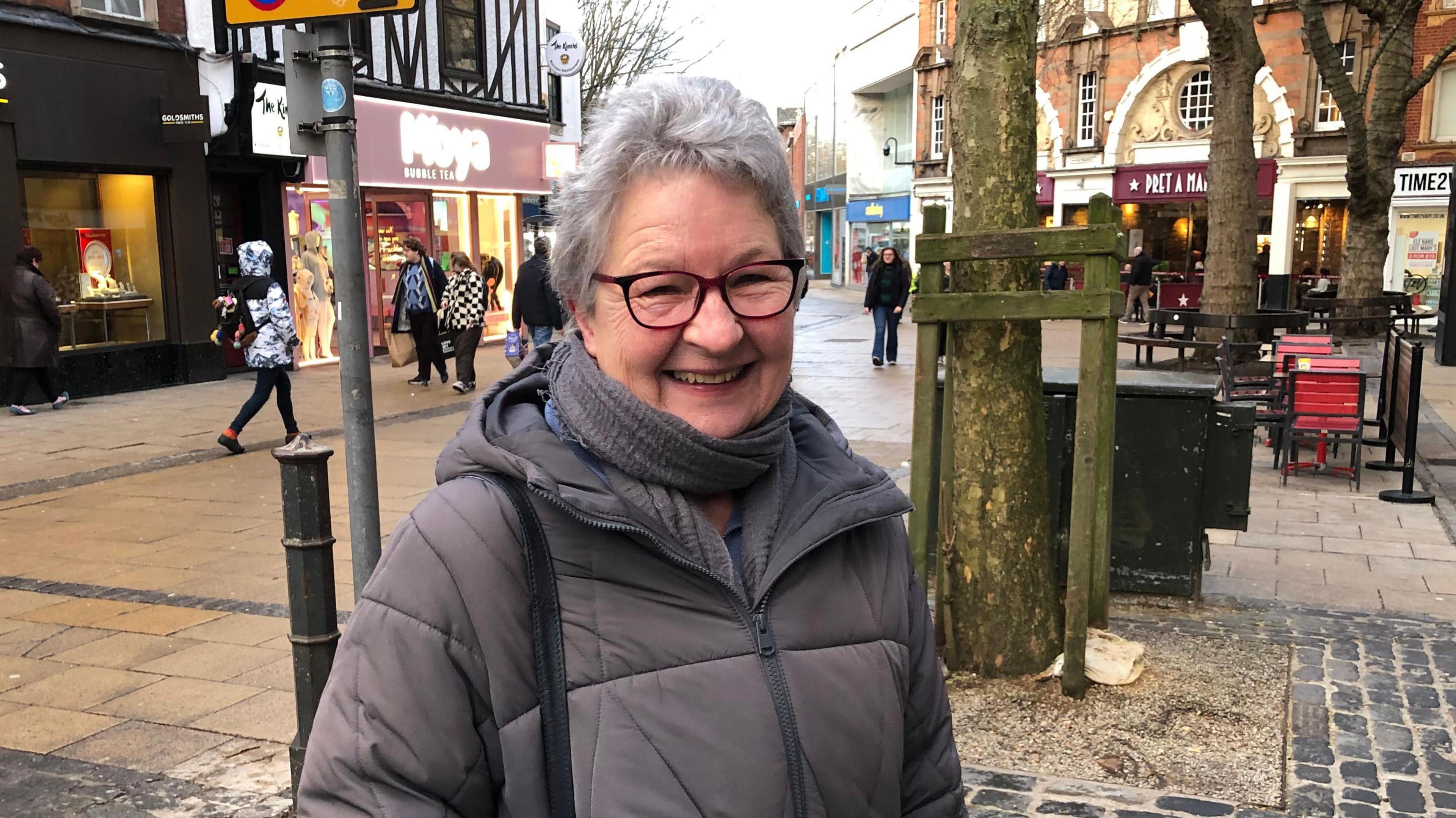 Margaret Duffield smiling in Norwich city centre. She's wearing a grey coat and red-rimmed glasses.