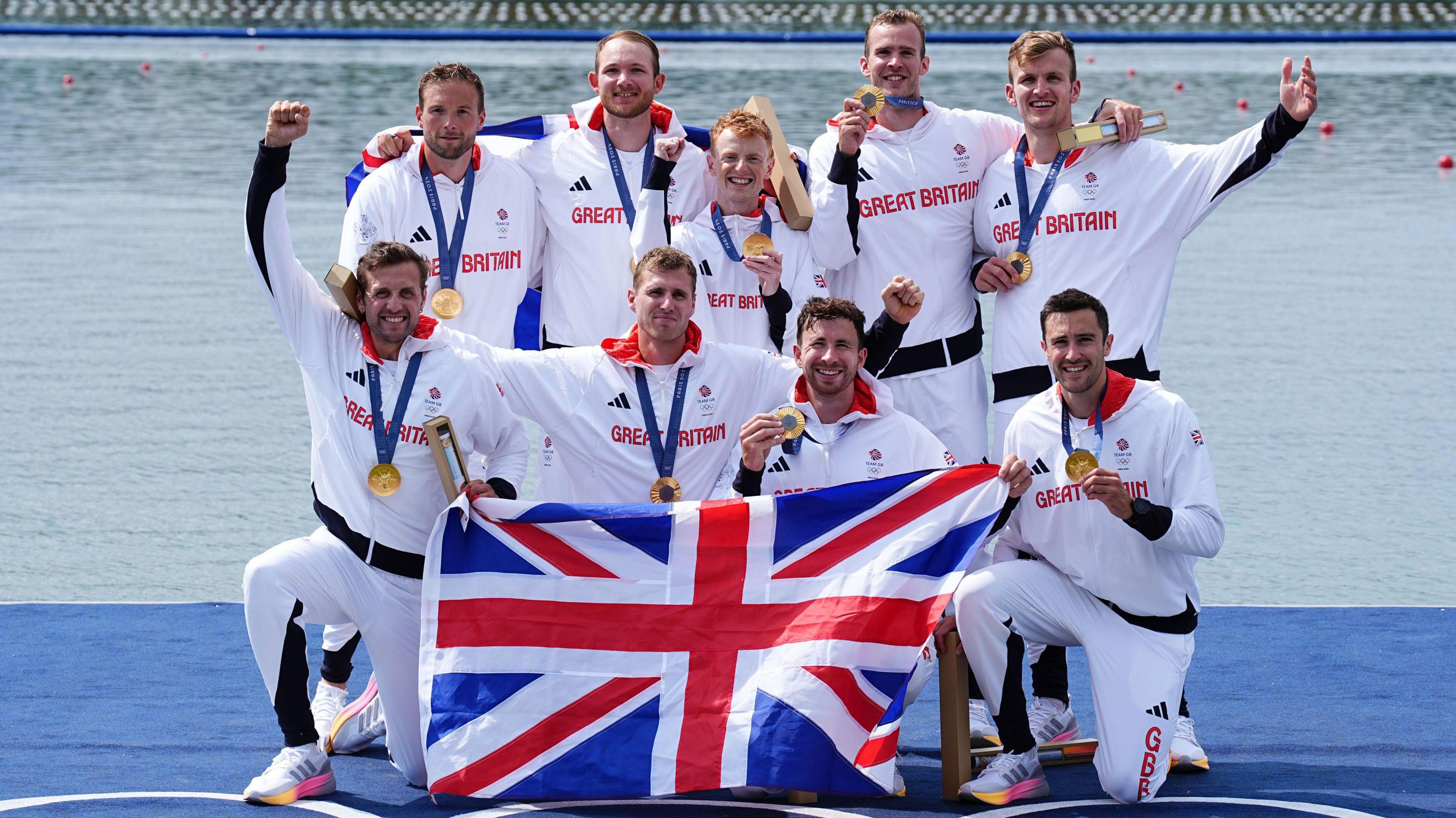 A team of nine men standing at the water's edge, wearing white Great Britain kits and holding a union flag. They are all smiling at the camera and holding up their gold medals.