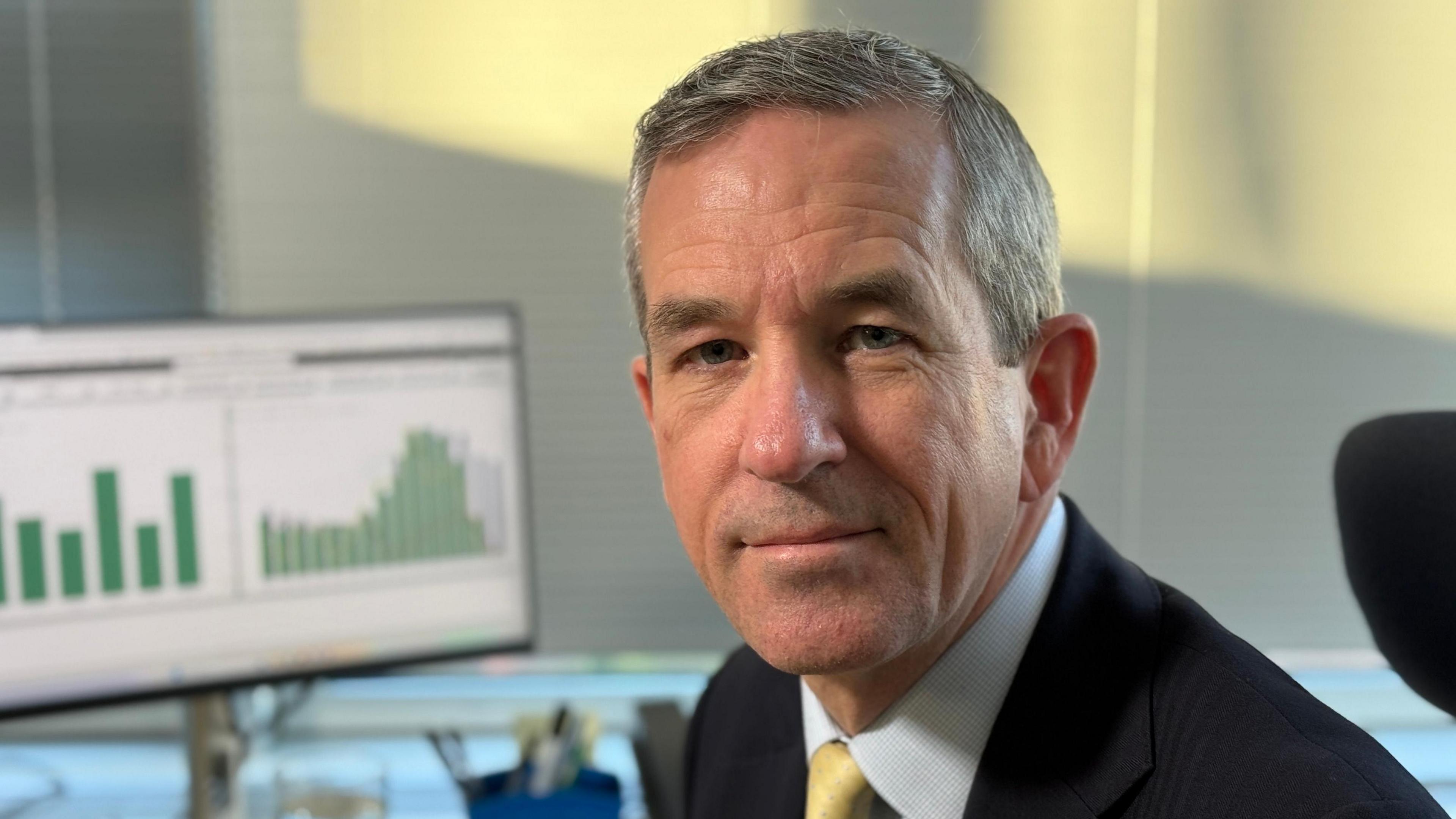 Neill Moloney smiles at the camera while sitting at a desk. He has short grey hair and is wearing a navy suit with a white shirt underneath and gold coloured tie. A computer screen can be seen to his right and is displaying two different graphs.