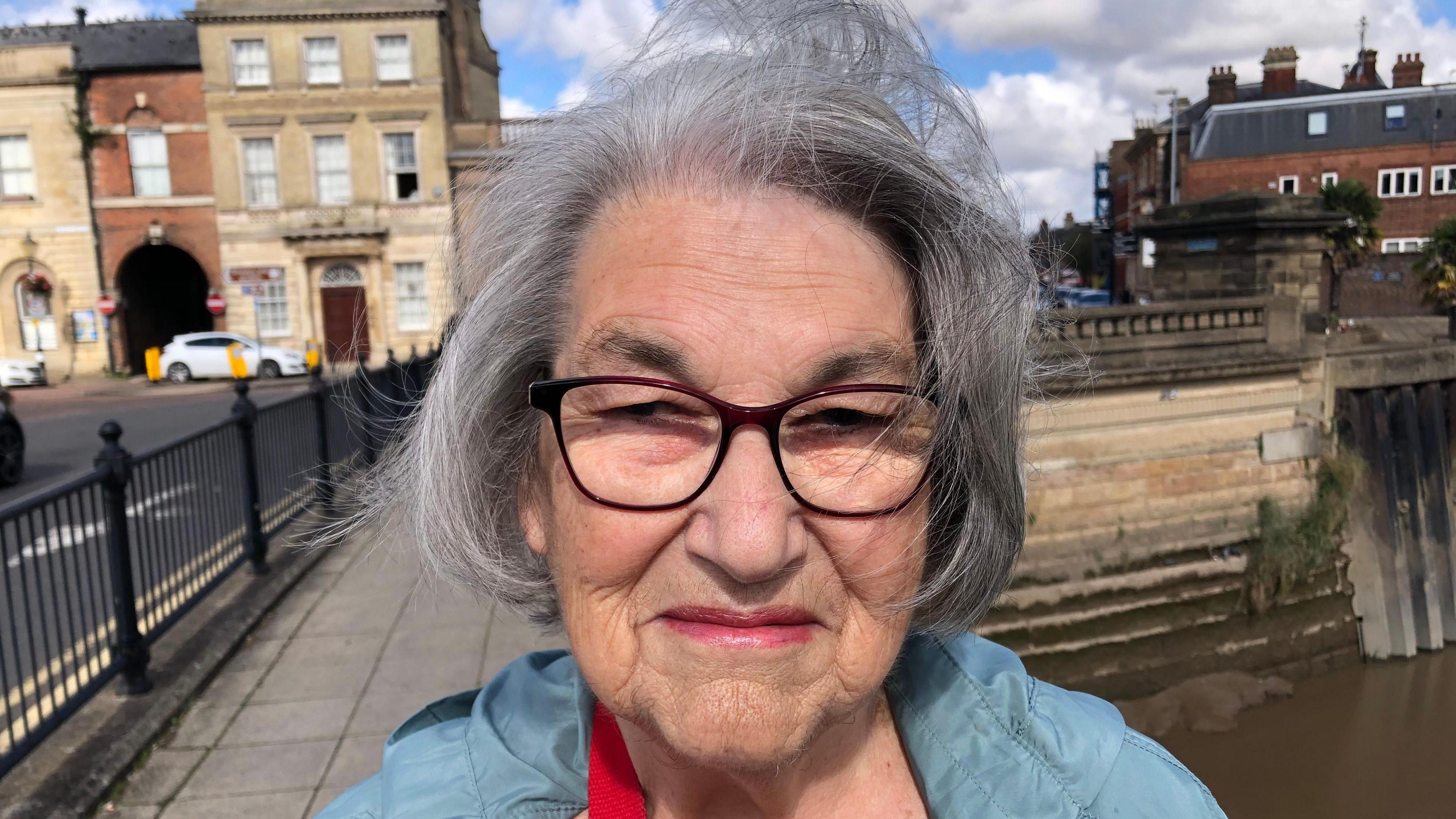 Bridget Holmes is photographed standing on a bridge smiling at the camera. She is a wearing a blue anorak and red glasses. 