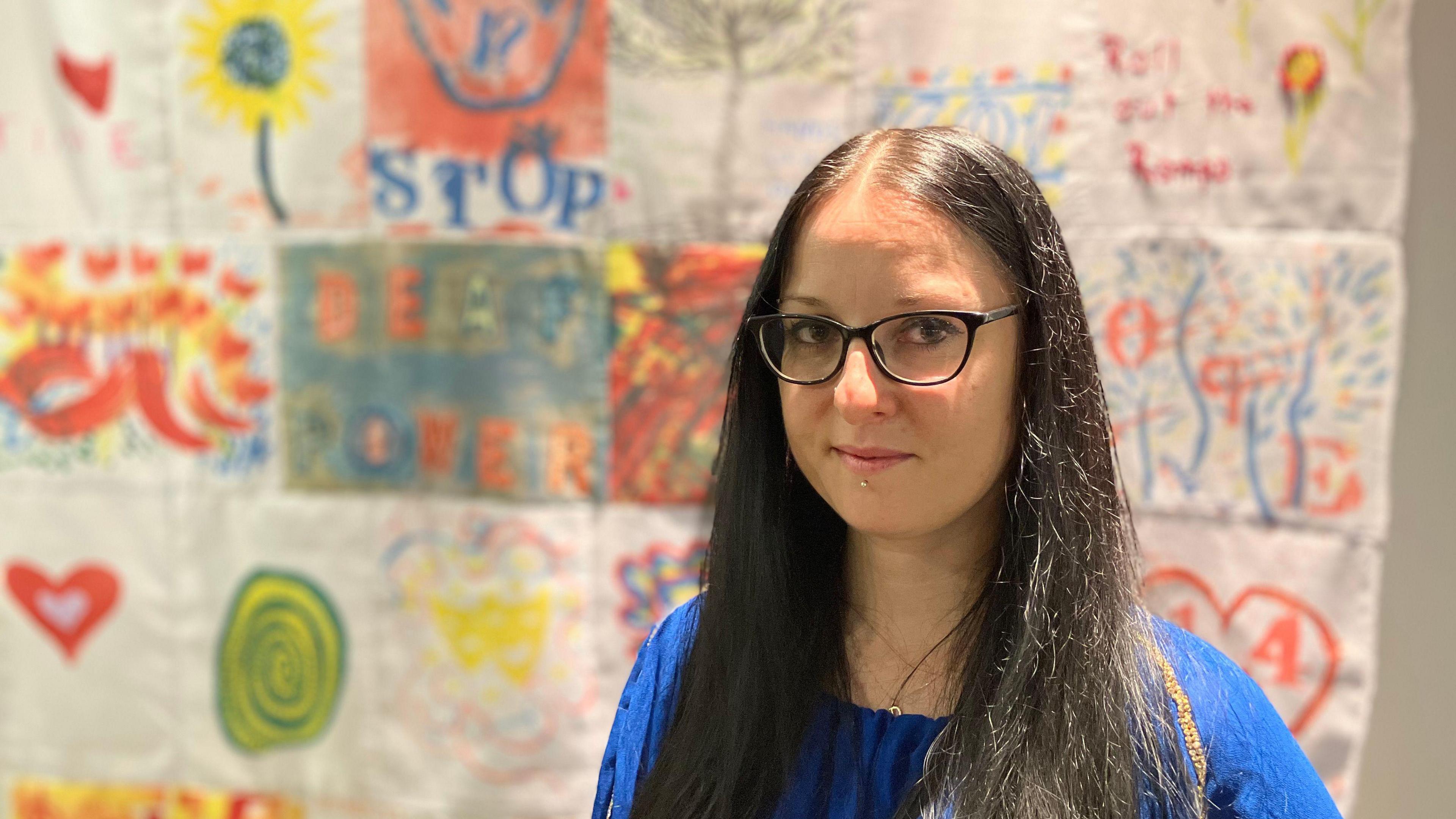 Dorata wearing a dark blue top and glasses, smiling for the camera in front of her white and coloured blanket on the wall