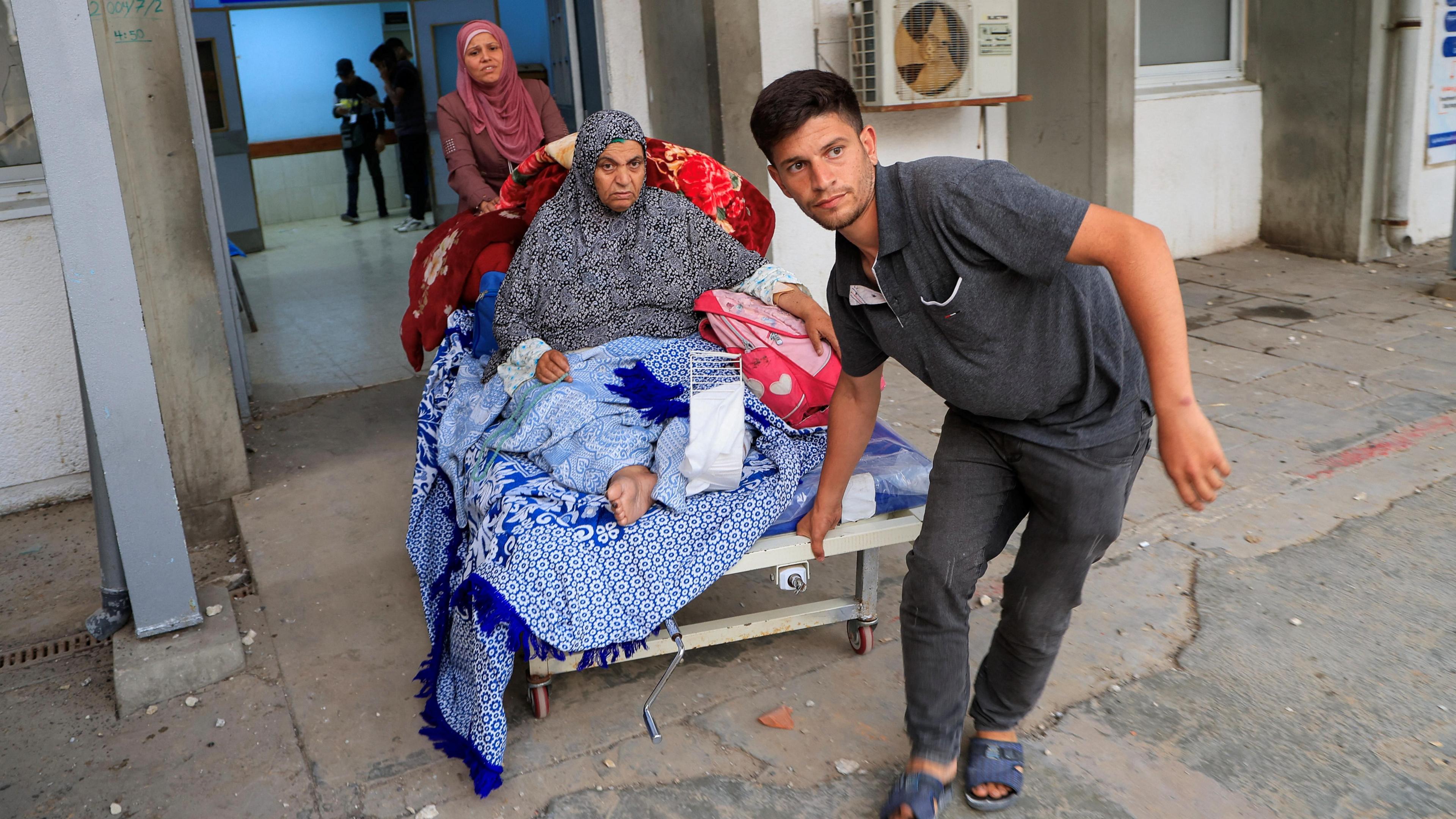 A Palestinian woman patient is evacuated from European Hospital. She is pictured sitting up in a bed on wheels, barefoot and covered in blankets. A younger man is pulling the bed out of a building, while another woman pushes it from behind.
