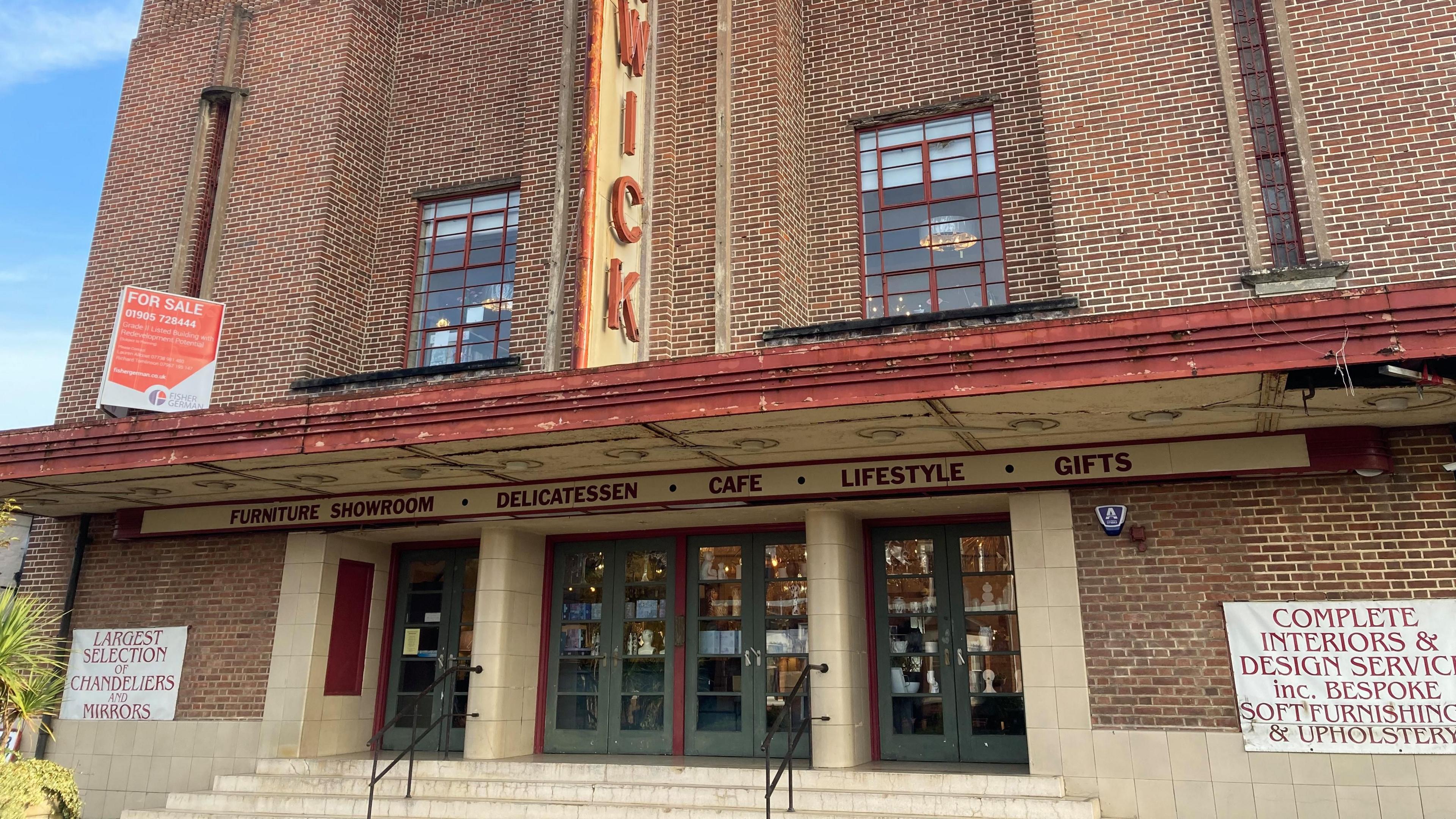 An art deco building with steps leading up to green front doors which are under a sign reading: "Furniture showroom - delicatessen  - cafe - lifestyle - gifts". There is a "for sale" sign on the side of the building.