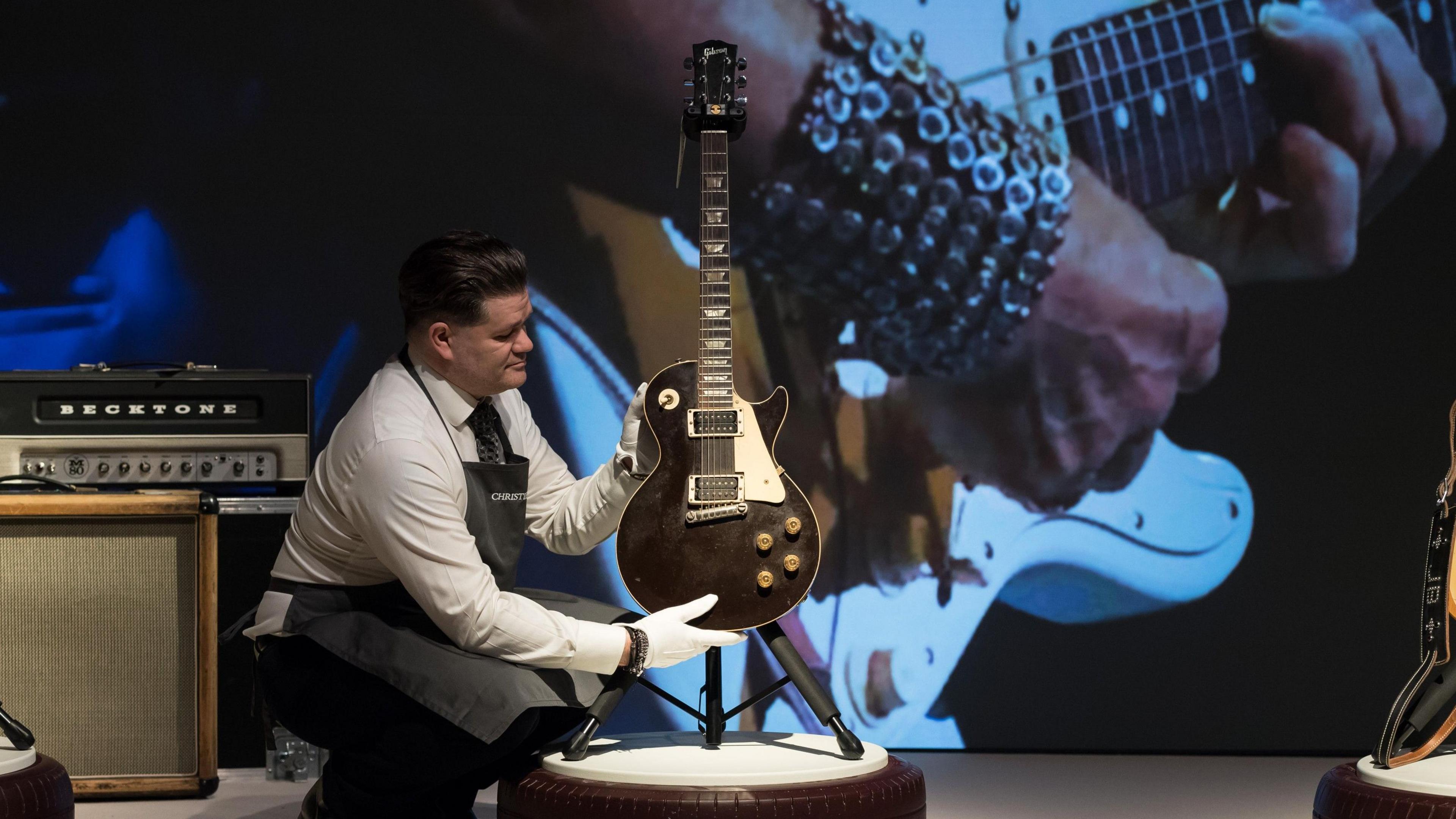 An auction house employee, wearing a white shirt, dark trousers and tie and a Christie's apron and white cotton gloves, holds Jeff Beck's famous Oxblood Les Paul guitar at the auction house, in front of a giant picture of the musician.