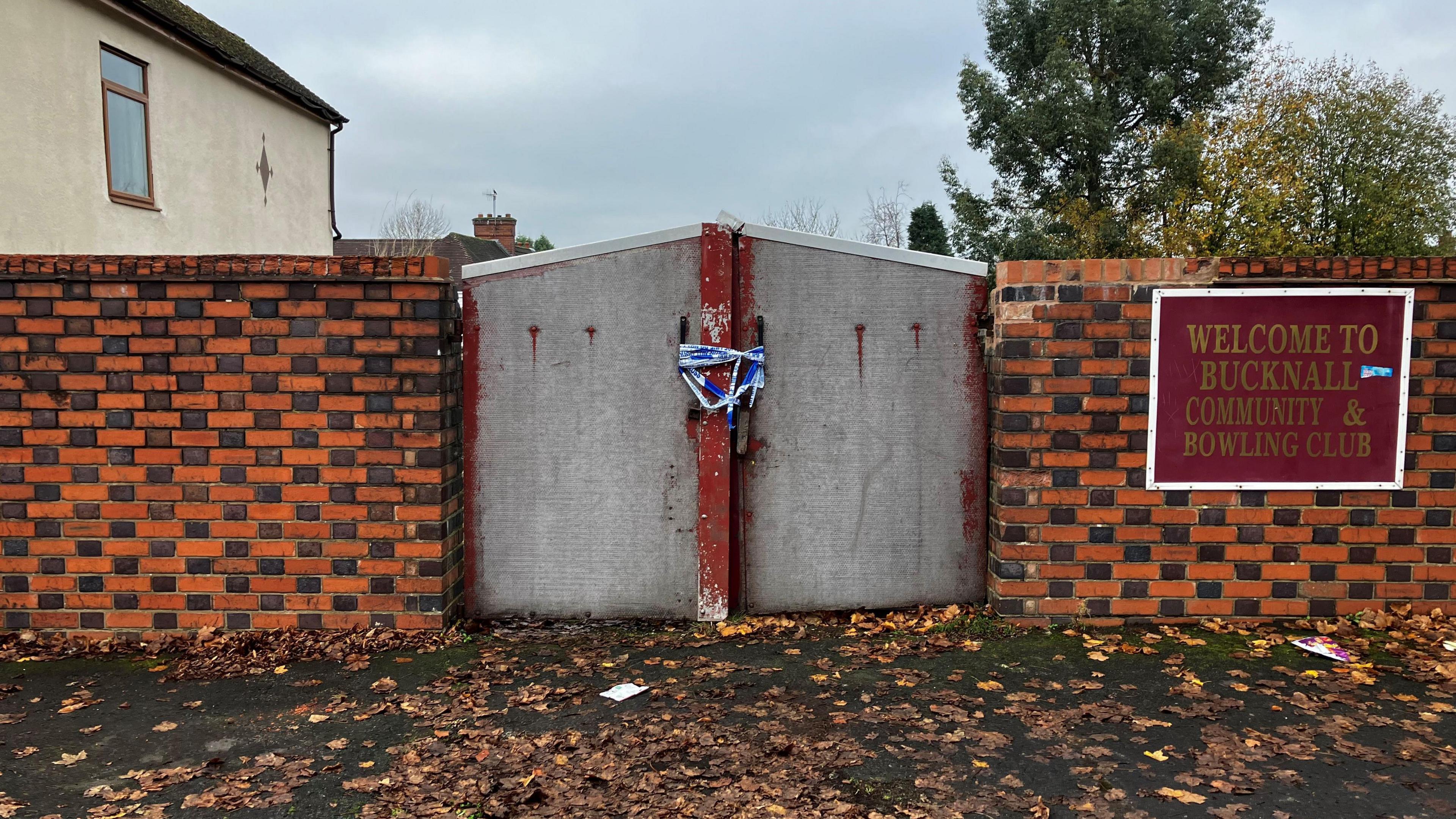 A metal gate, set in a red and grey brick checkerboard wall. There is a sign which reads 'welcome to Bucknall community and bowling club'.