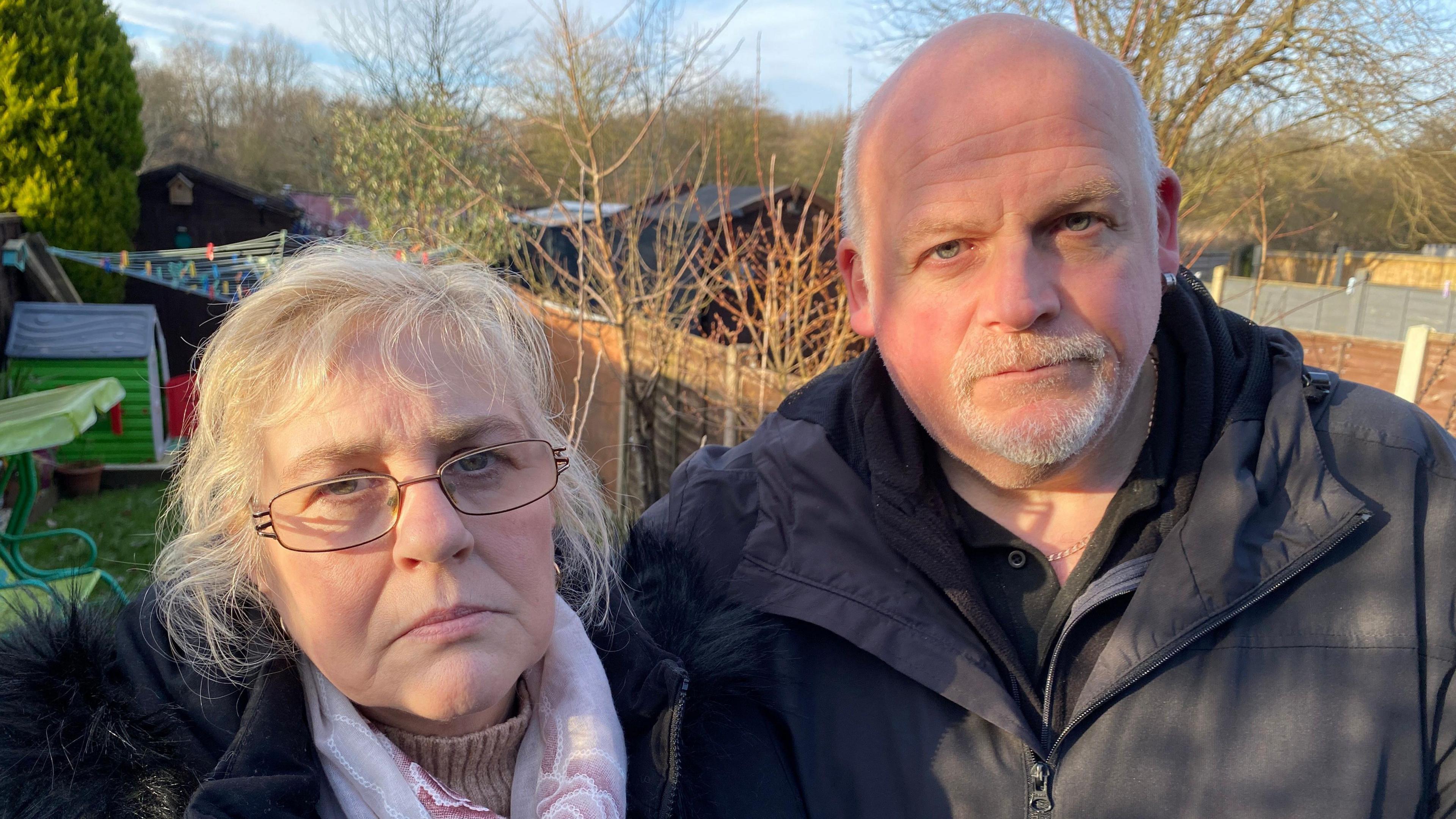 Amanda Newton and Phil Shakespeare standing side by side. They are in a back garden, and a clothes line can be seen behind them. It is winter time and they both have coats on. They look unhappy. 