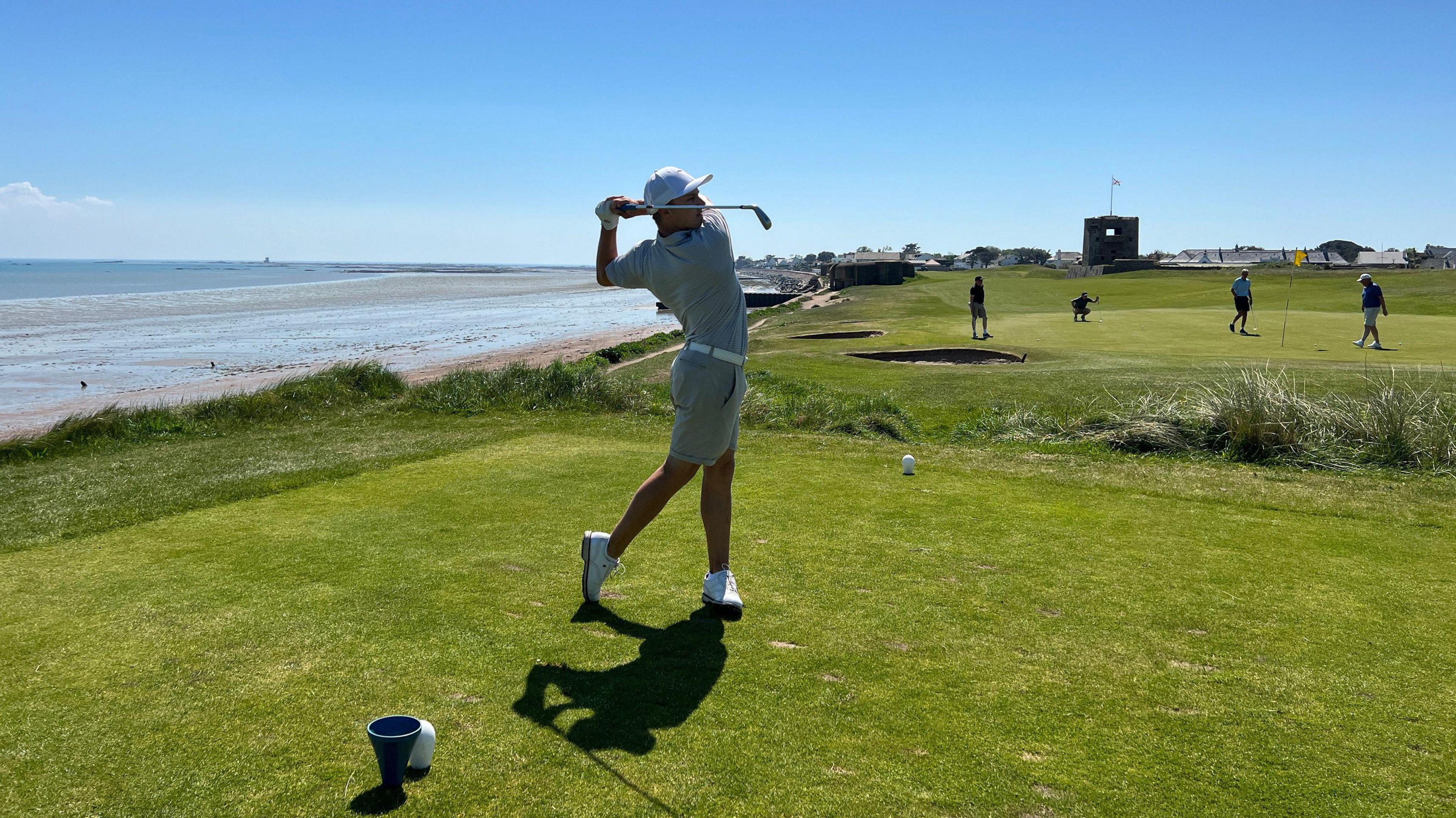 Josef has swung his golf club on a green with others playing the background and to the left there is a beach and seaside. Josef is wearing white shoes, cream shorts and t-shirt and a white cap.