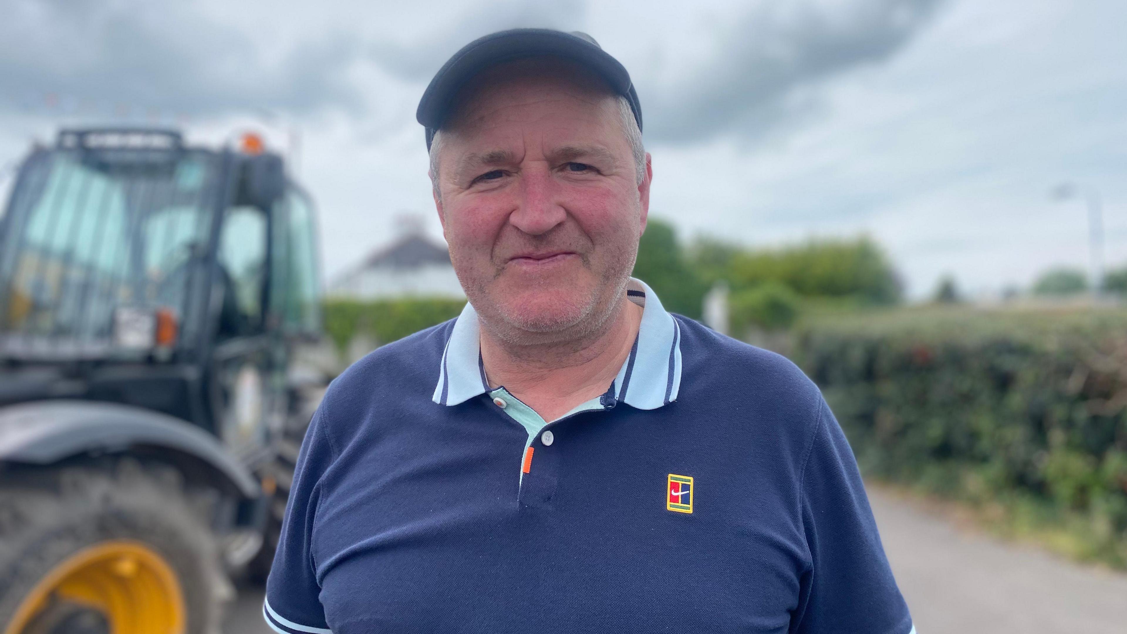 Mick is smiling into the camera. He is wearing a blue polo shirt with a red crest. The background is blurred, ut you can make out a tractor on a country road with yellow wheel rims.