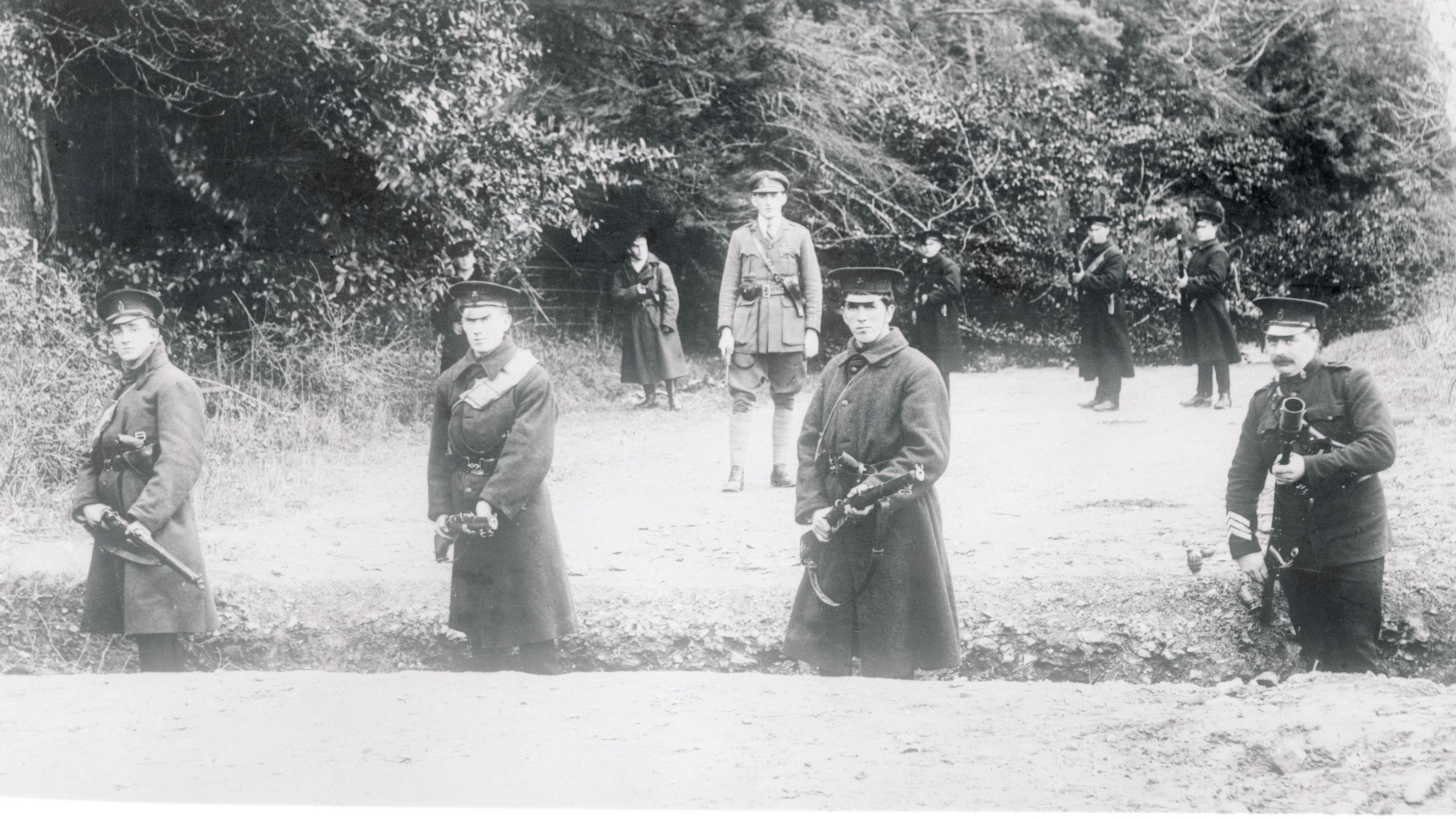A black and white archive photo of armed police guarding a rural border road in Northern Ireland circa 1922.  Four officers in peaked caps and coats are standing in a shallow trench in the foreground, pointing guns.  More officers are standing guard in the background. 