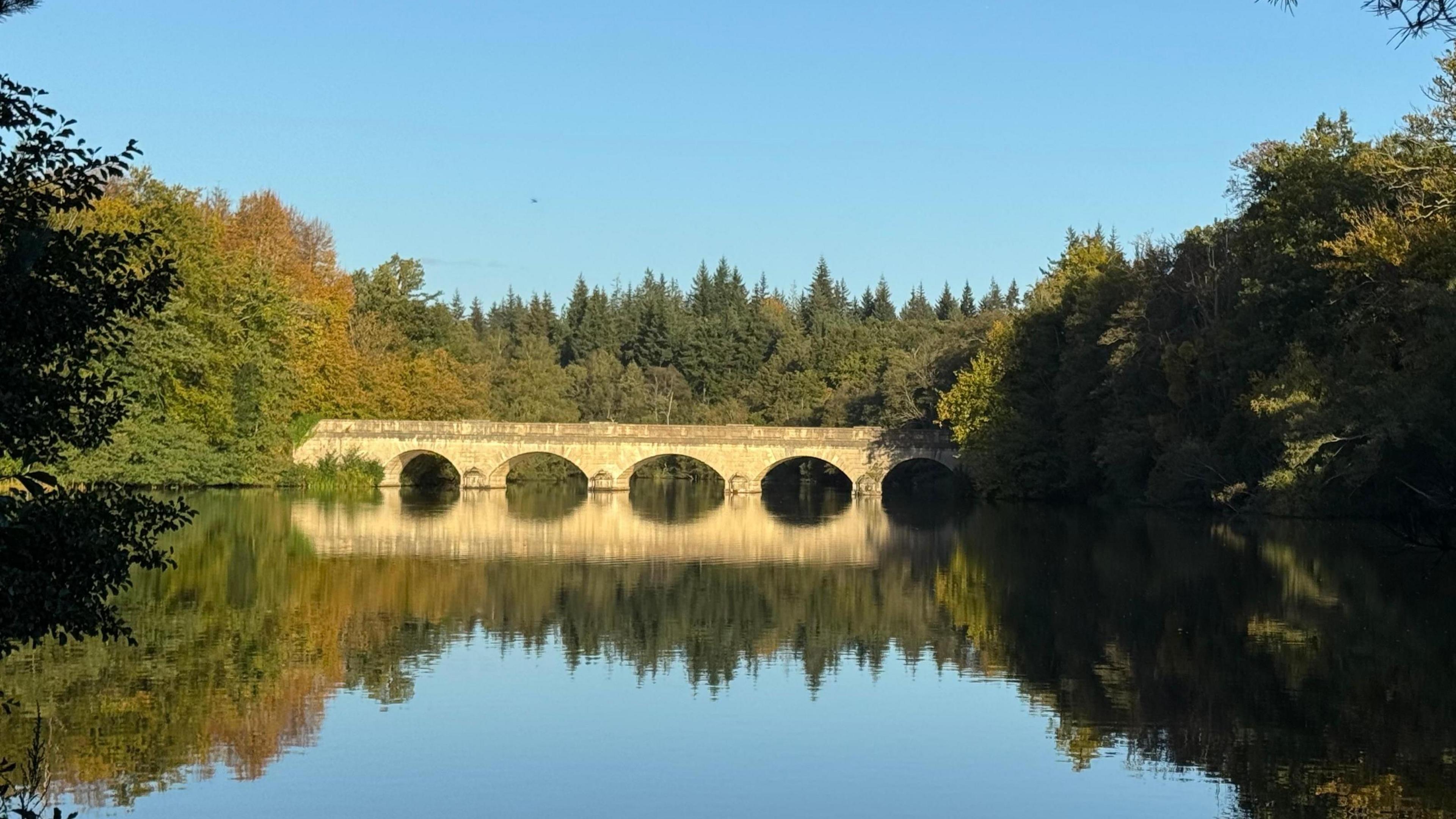 A yellow stone bridge basks in brought sunshine as it spans over a lake with its reflection perfectly replicated in the smooth surface of the water. Trees line either side and a cloudless sky is a bright blue above