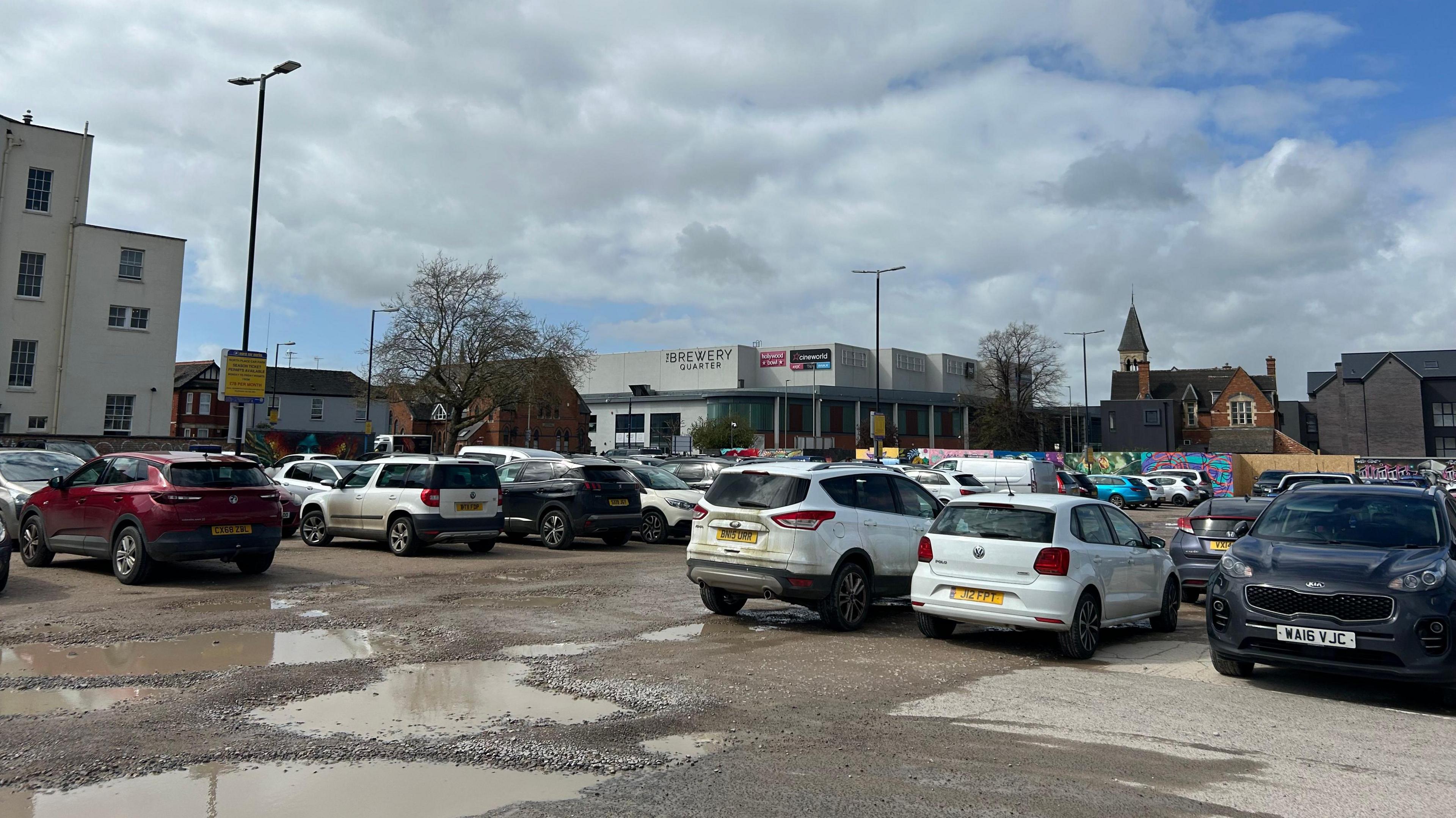 Cars parked in a car park with a wet surface, and water filling the potholes, and a shopping centre in the background