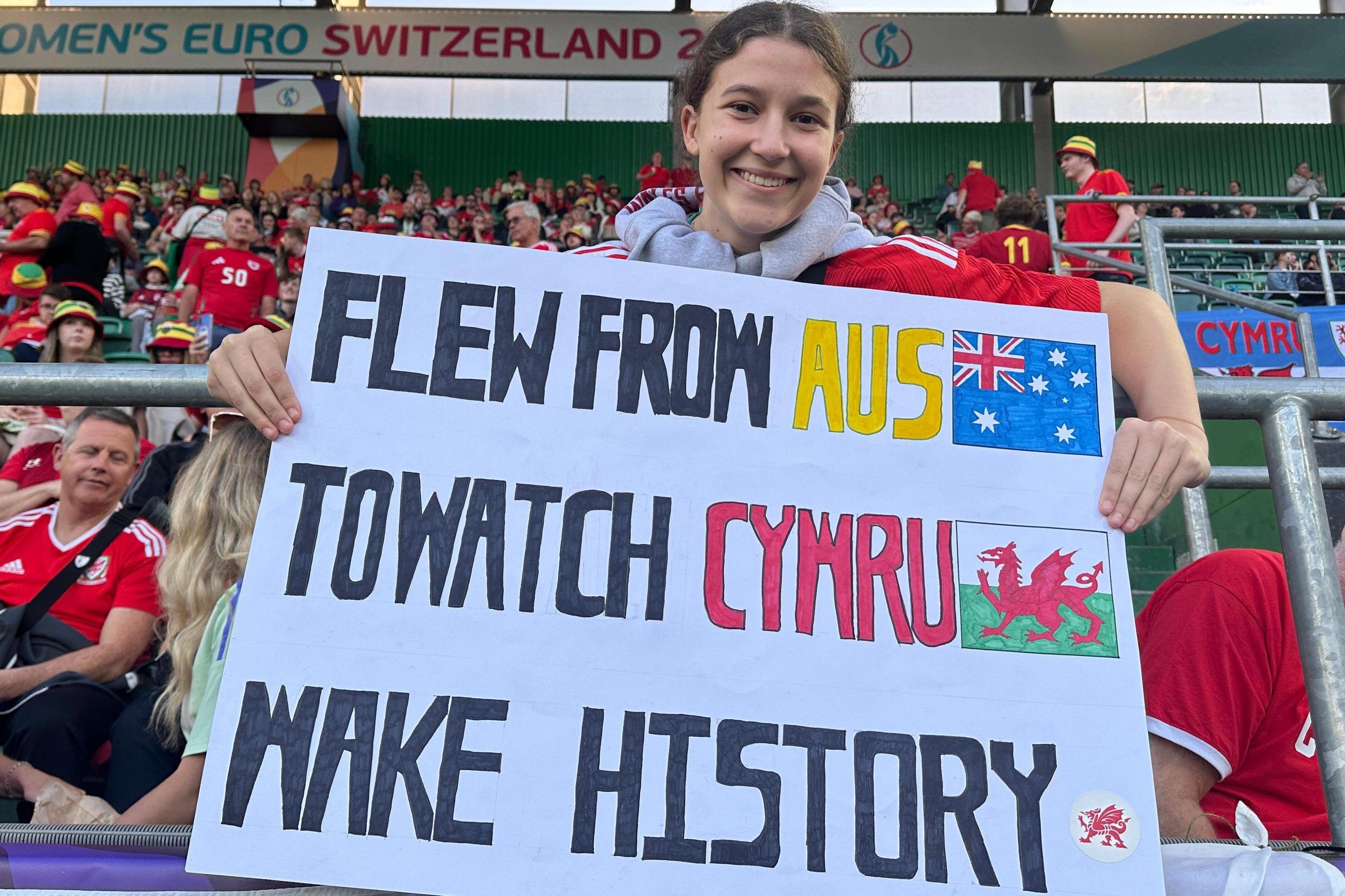 Helena is smiling at the camera while clutching a banner that read ‘Flew from Aus to watch Cymru make history’. It is hand drawn with pictures of the Australian flag beside the word 'Aus' and the Welsh flag beside the word 'Cymru'. She is wearing a red shirt with a grey jumper over it. She is sitting in the crowd of the stands.