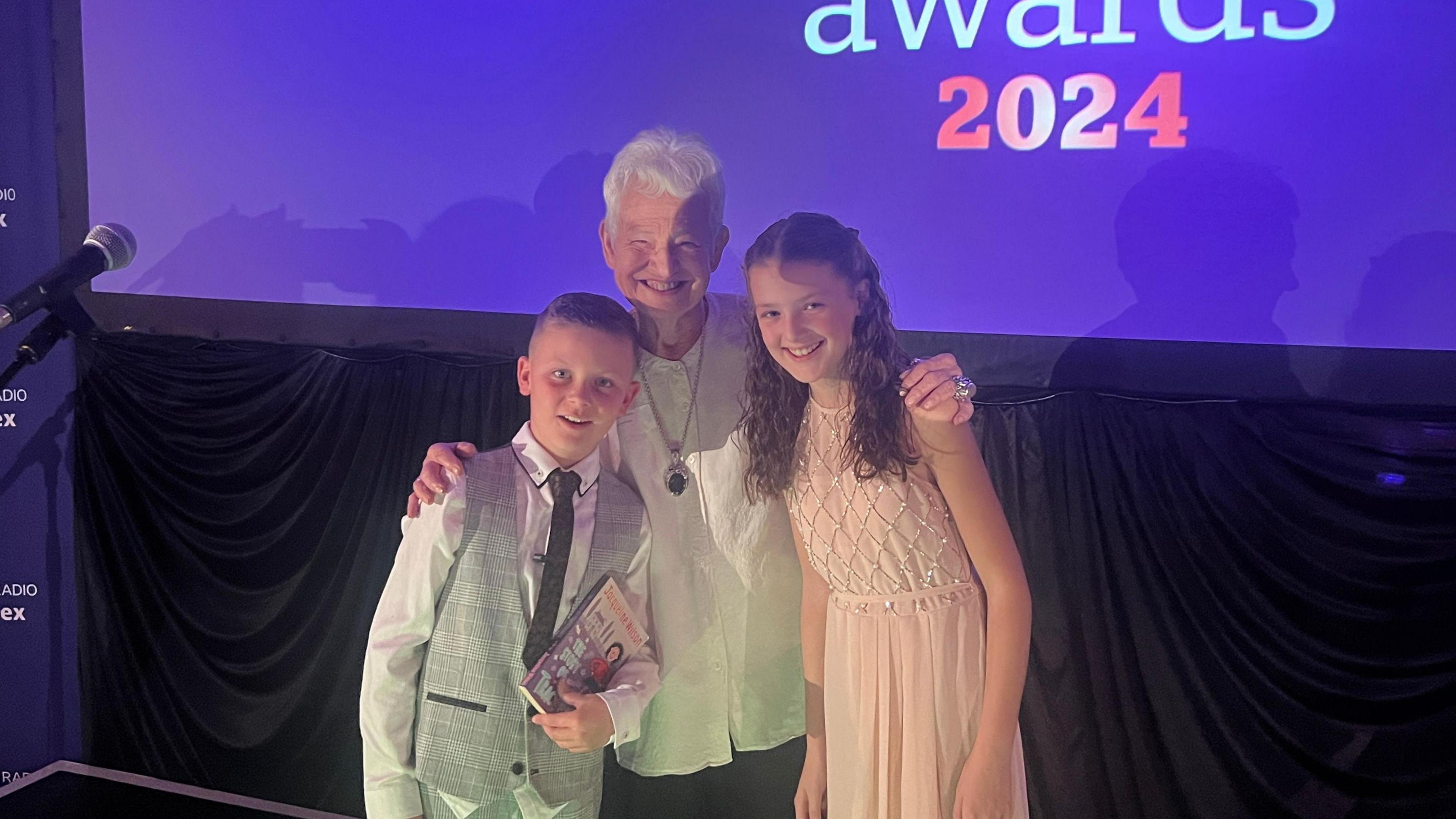 Dame Jacqueline Wilson with two young fans Hayden and Sennen Calver on the stage with a purple backdrop