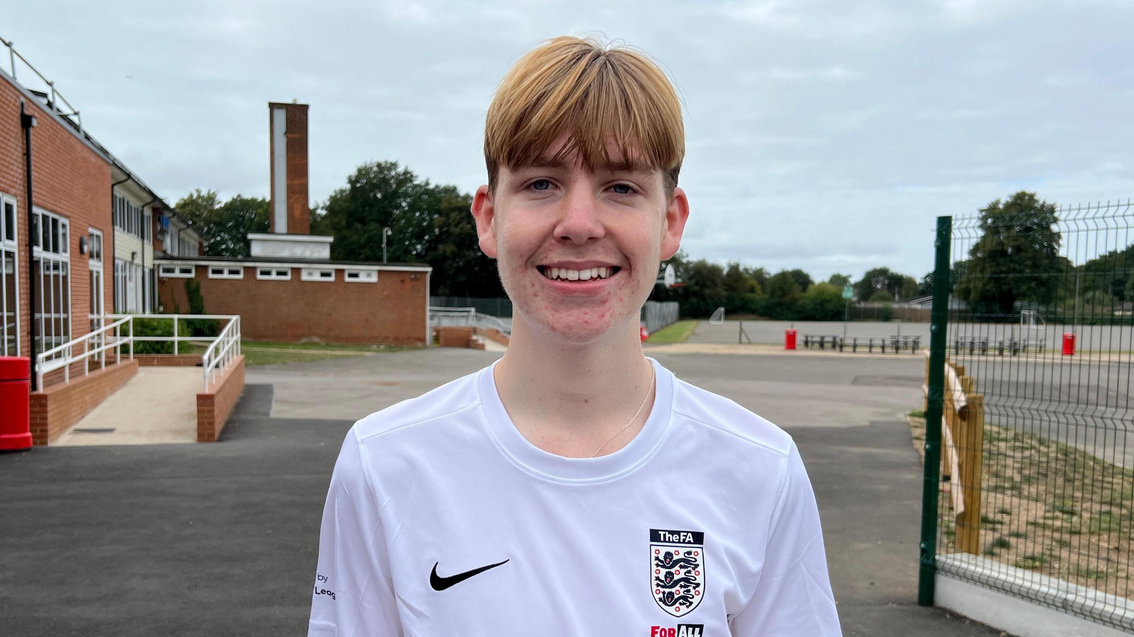 George is standing outside in the school grounds, looking at the camera and smiling. He has short red hair, with a long fringe. He is wearing a white round neck football t shirt. Behind him is a school building to the left and an empty car park.