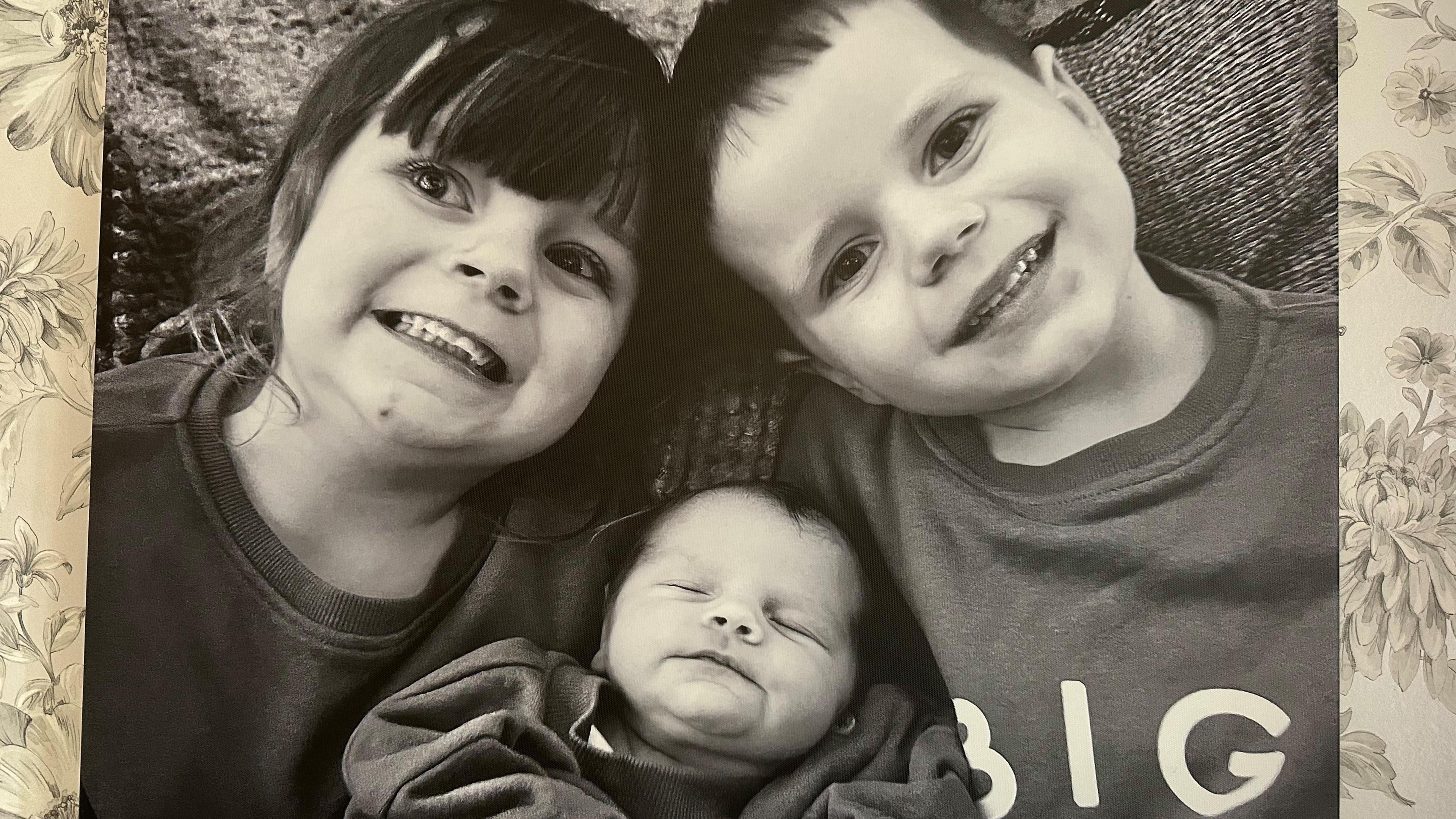 A black and white photo of a boy and girl smiling with a baby cuddled between them. The photo is hung on a wallpapered wall