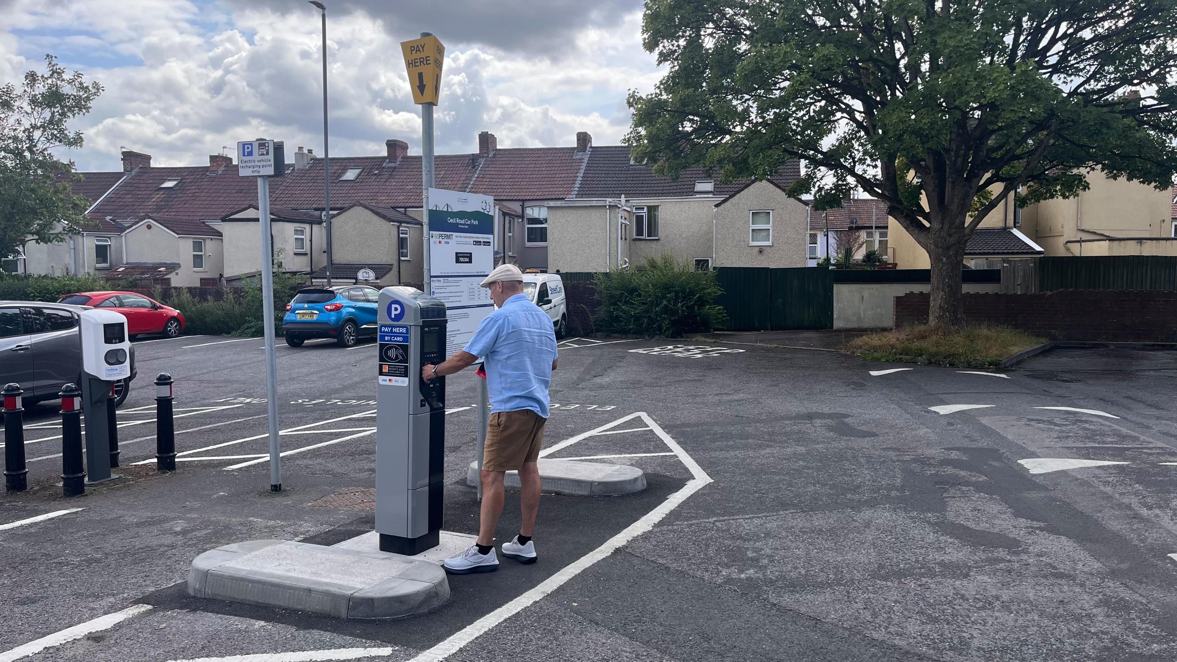 A man wearing a blue shirt, brown shorts and a cream flat cap pays for his parking in a Kingswood car park. Three cars can be seen parked in the background with a line of houses in the distance. 