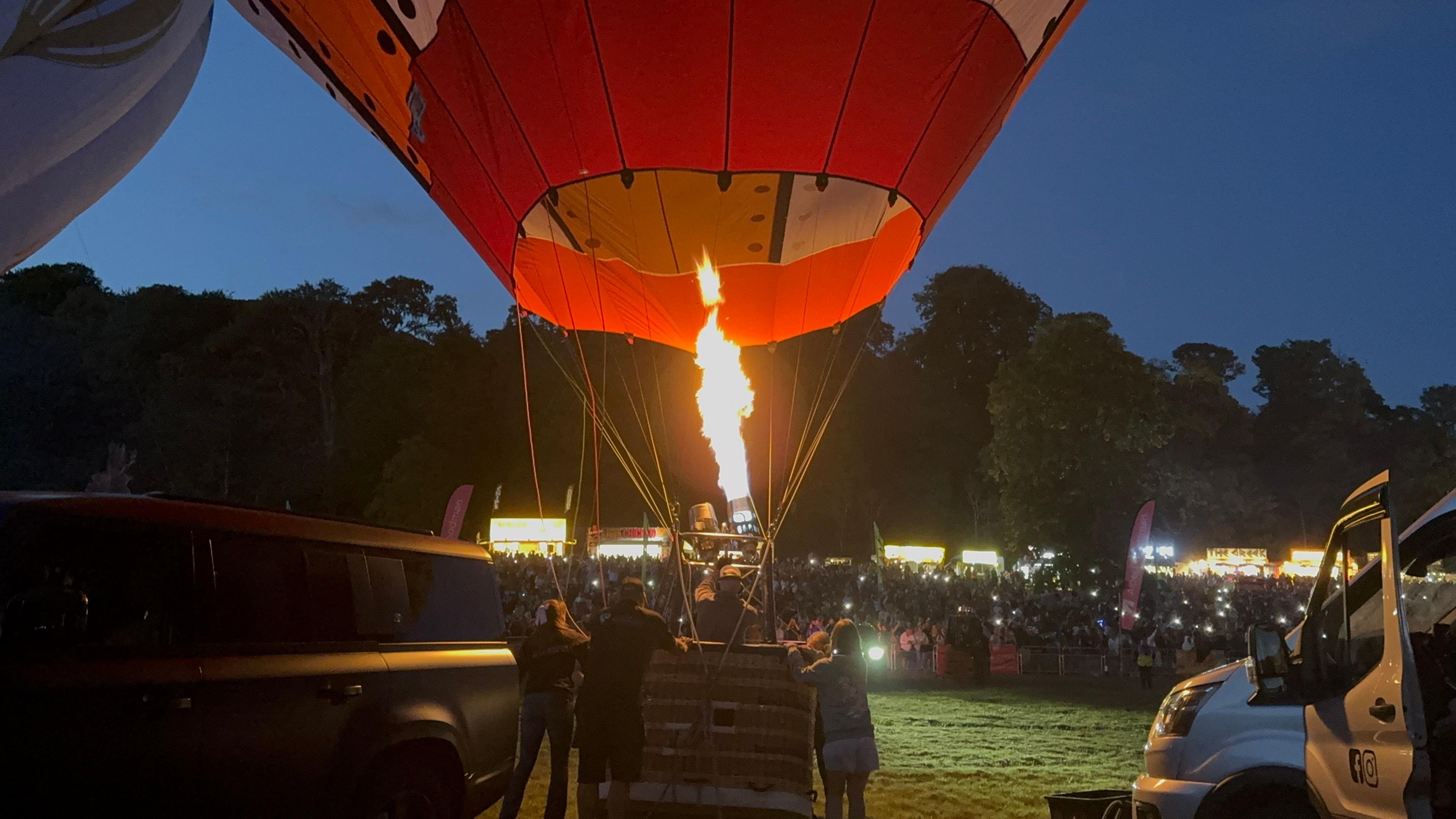 A group of people gathered around a hot air balloon basket. Their faces are lit up by a burners' flames in full force. They are surrounded by people with their phone lights, on filming the situation.