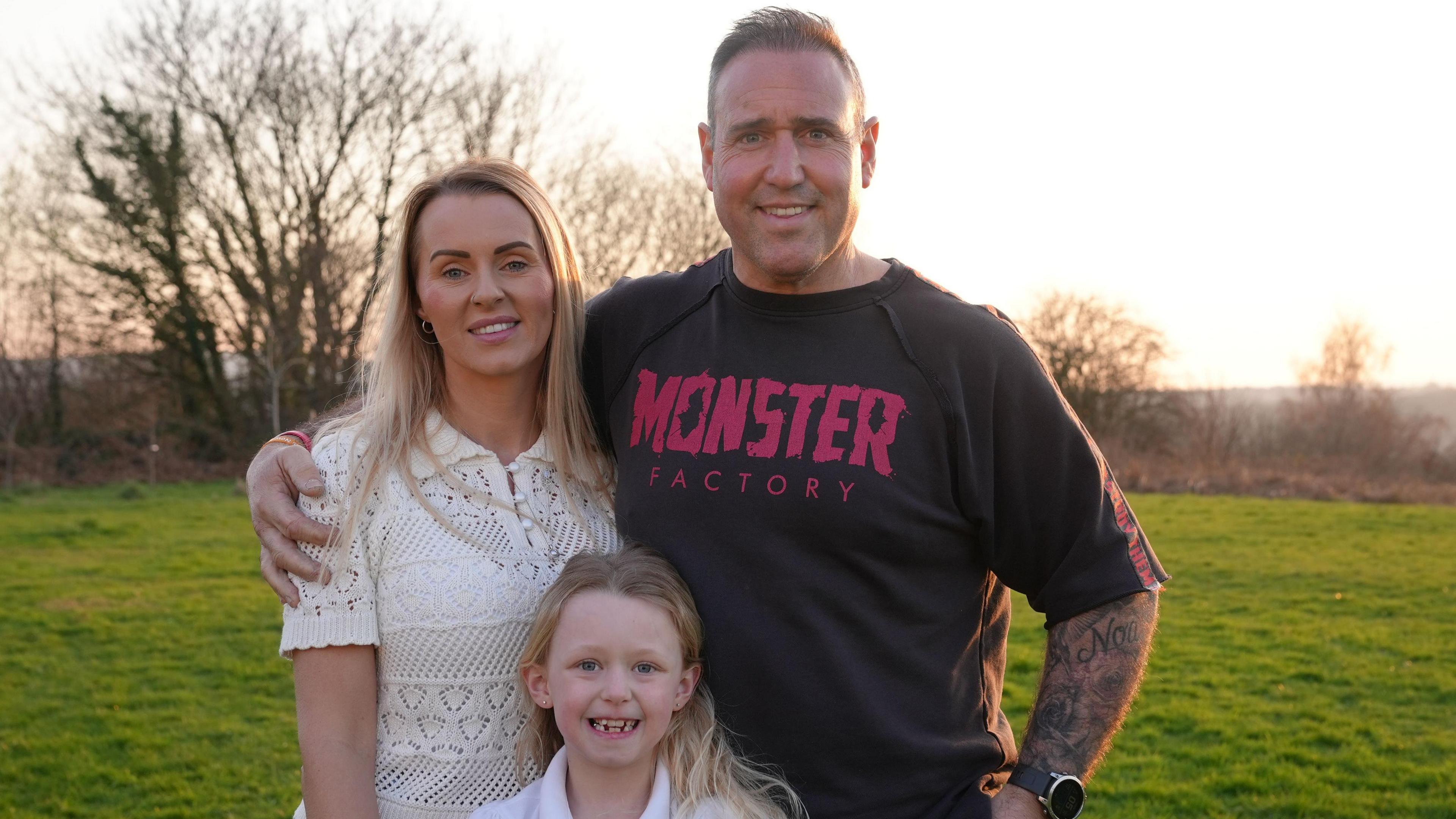 Emma, Scott a Noa-Rose yn sefyll ac yn gwenu gyda'i gilydd ar gae yn y machlud. 