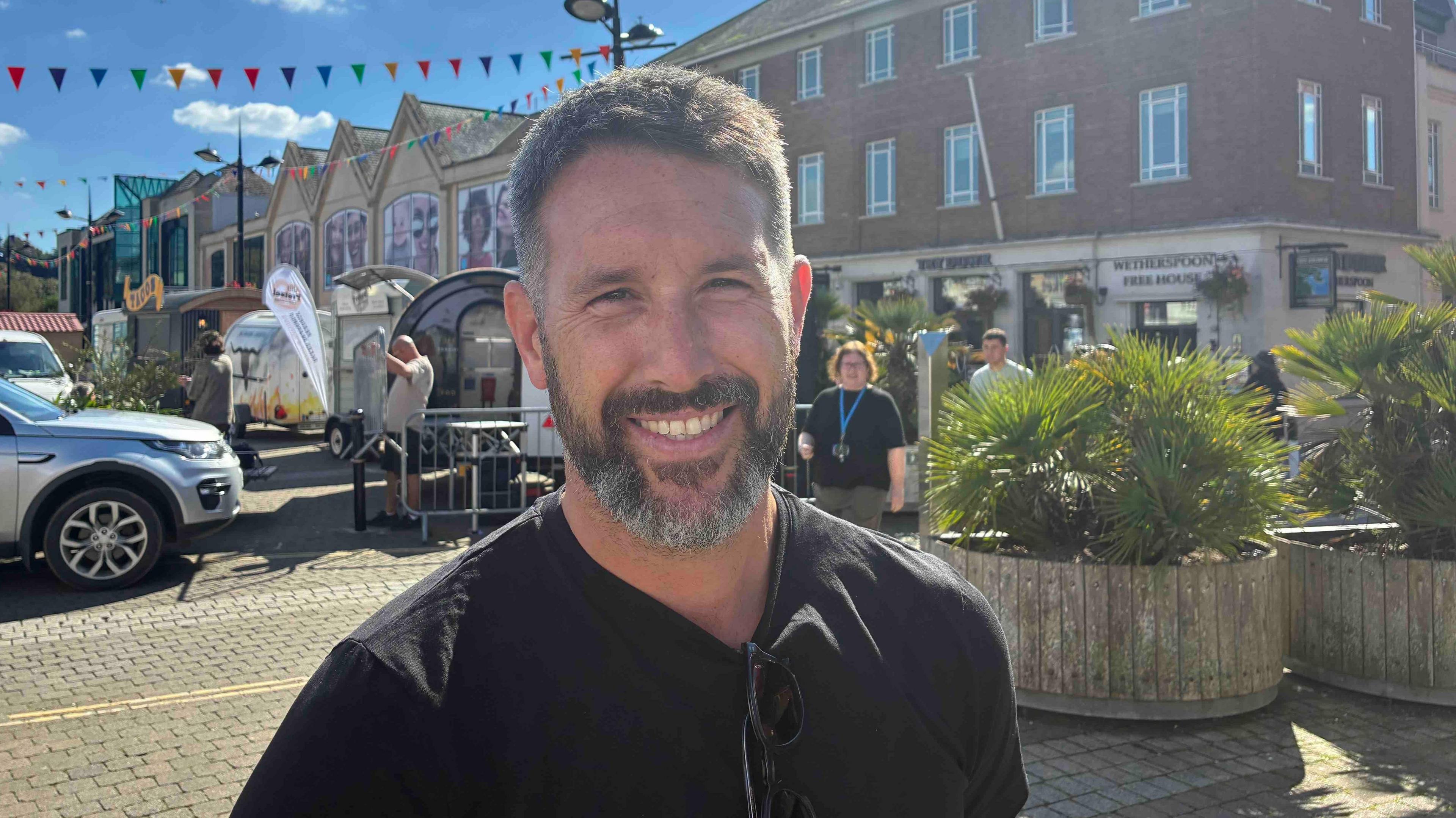 A smiling man is wearing a black top. He is standing near food stalls in Lemon Quay in Truro city centre, with food stalls behind him.
