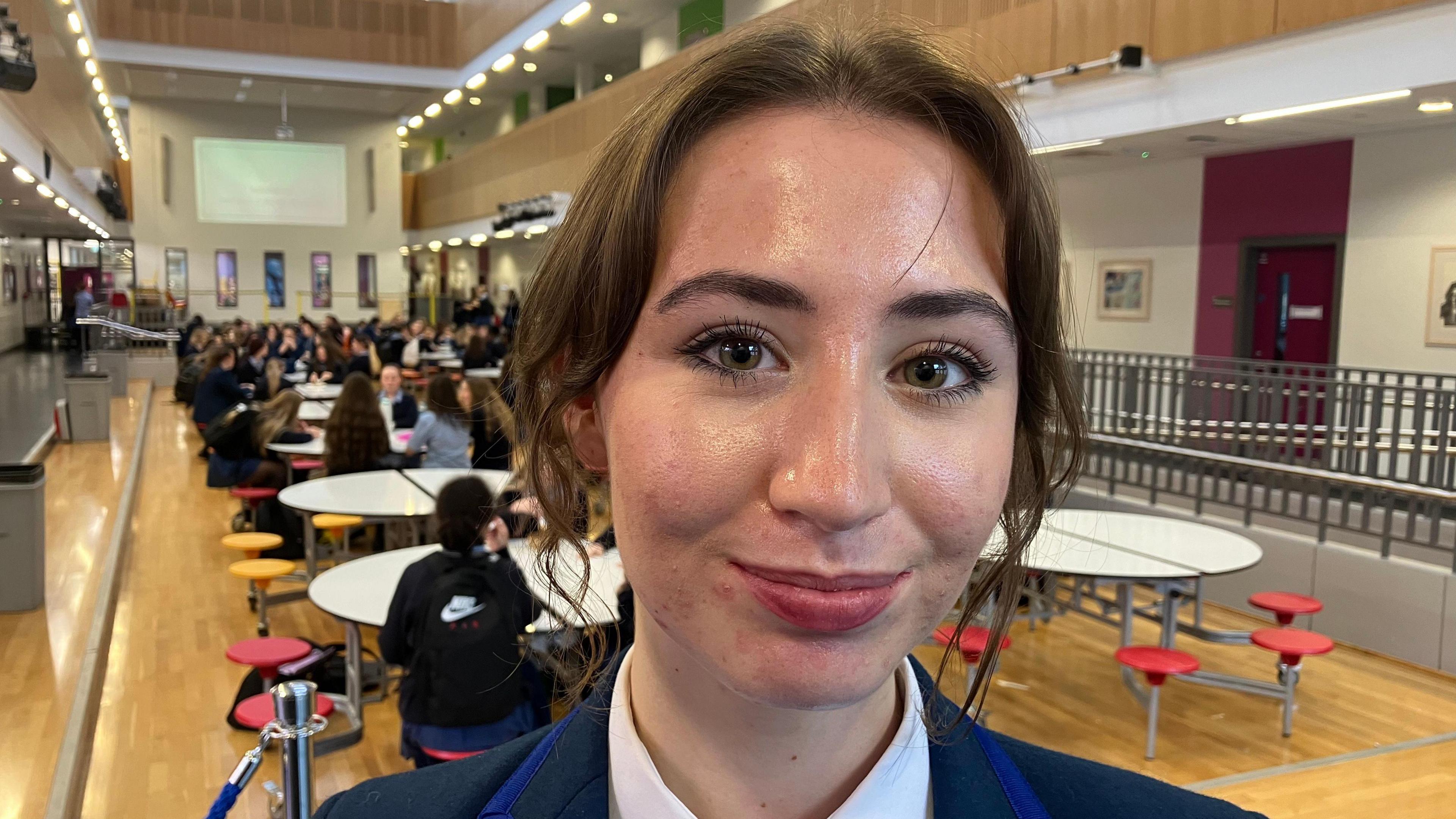Sophie. She has dark hair tied back with bits of hair down the side of her face. She is wearing a blue school blazer and white shirt. She is standing in a school canteen with pupils sitting at tables in the background.