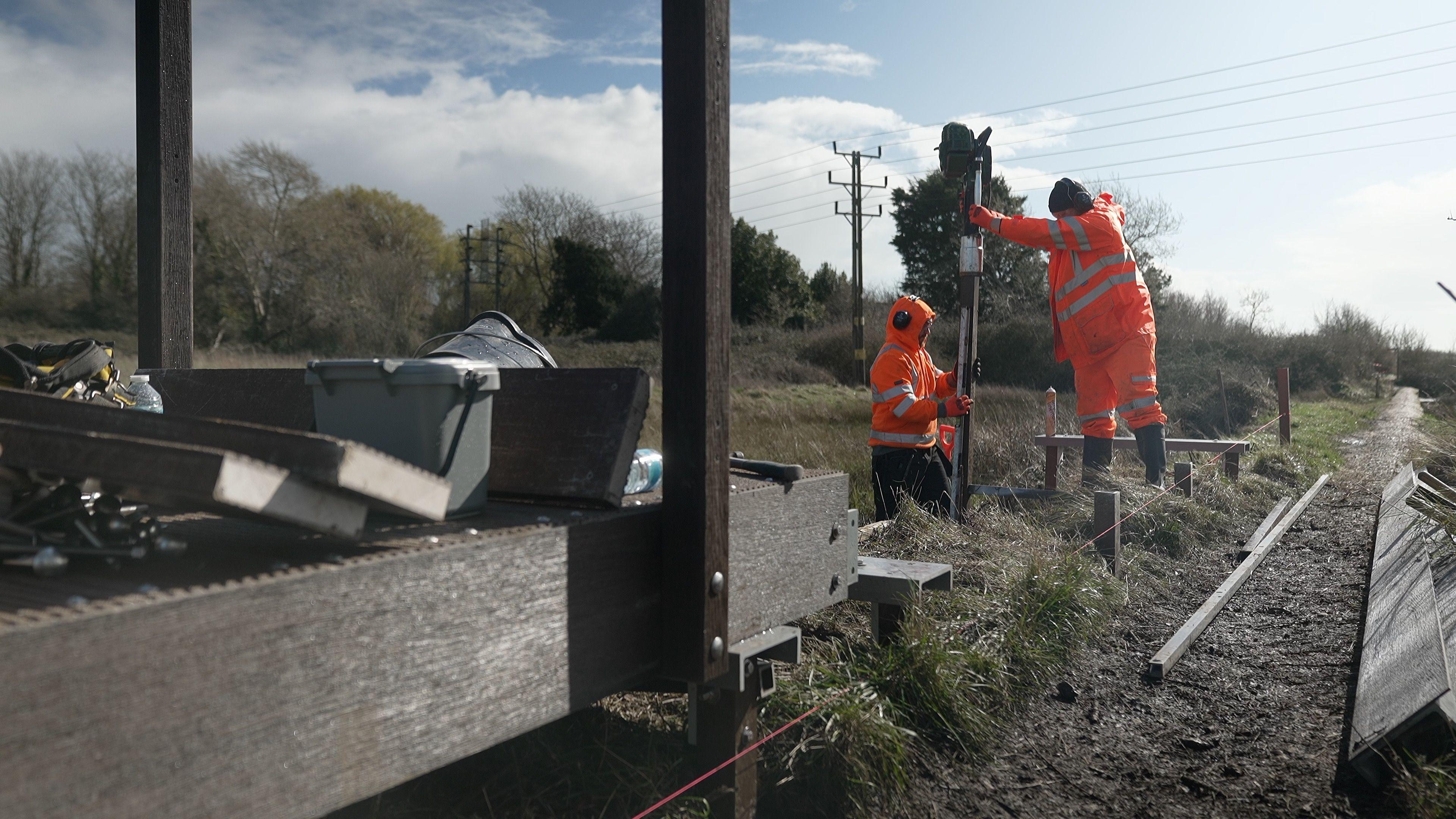 Two men in orange high-vis outfits and protective headsets are working on the path. They are standing with one of them holding a pile and the other one drilling the pile into the ground. In the foreground, a wooden walkway is seen and there are the men's tools on it.