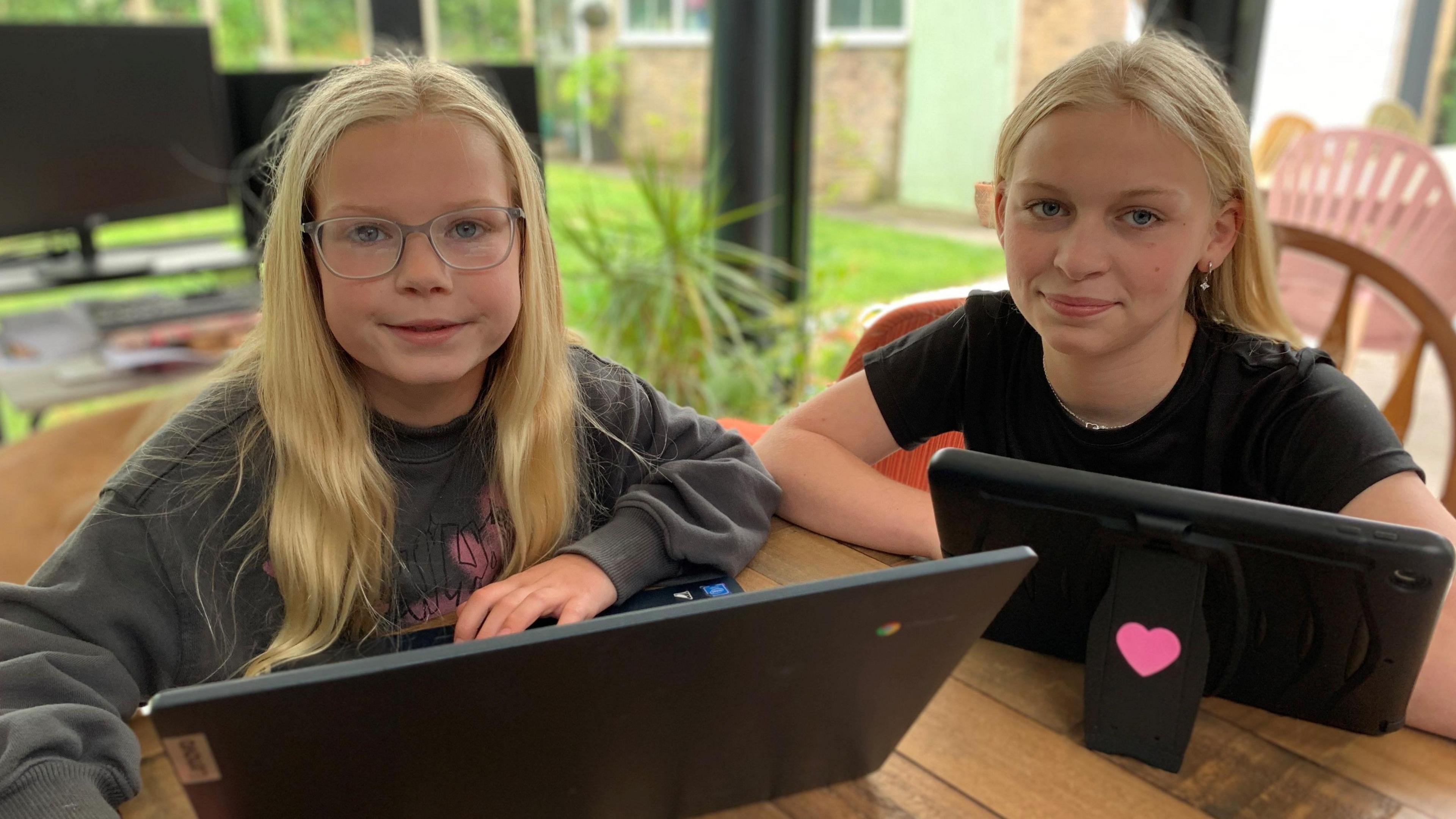 Rosa is an 8 year old girl with long blond hair and wearing glasses. She is wearing a grey jumper and smiling. On her righthand side is 14 year old sister Matilda with long blond hair, wearing silver ear rings and a black T-shirt. Both are sat at a table with devices in front of them and smiling.