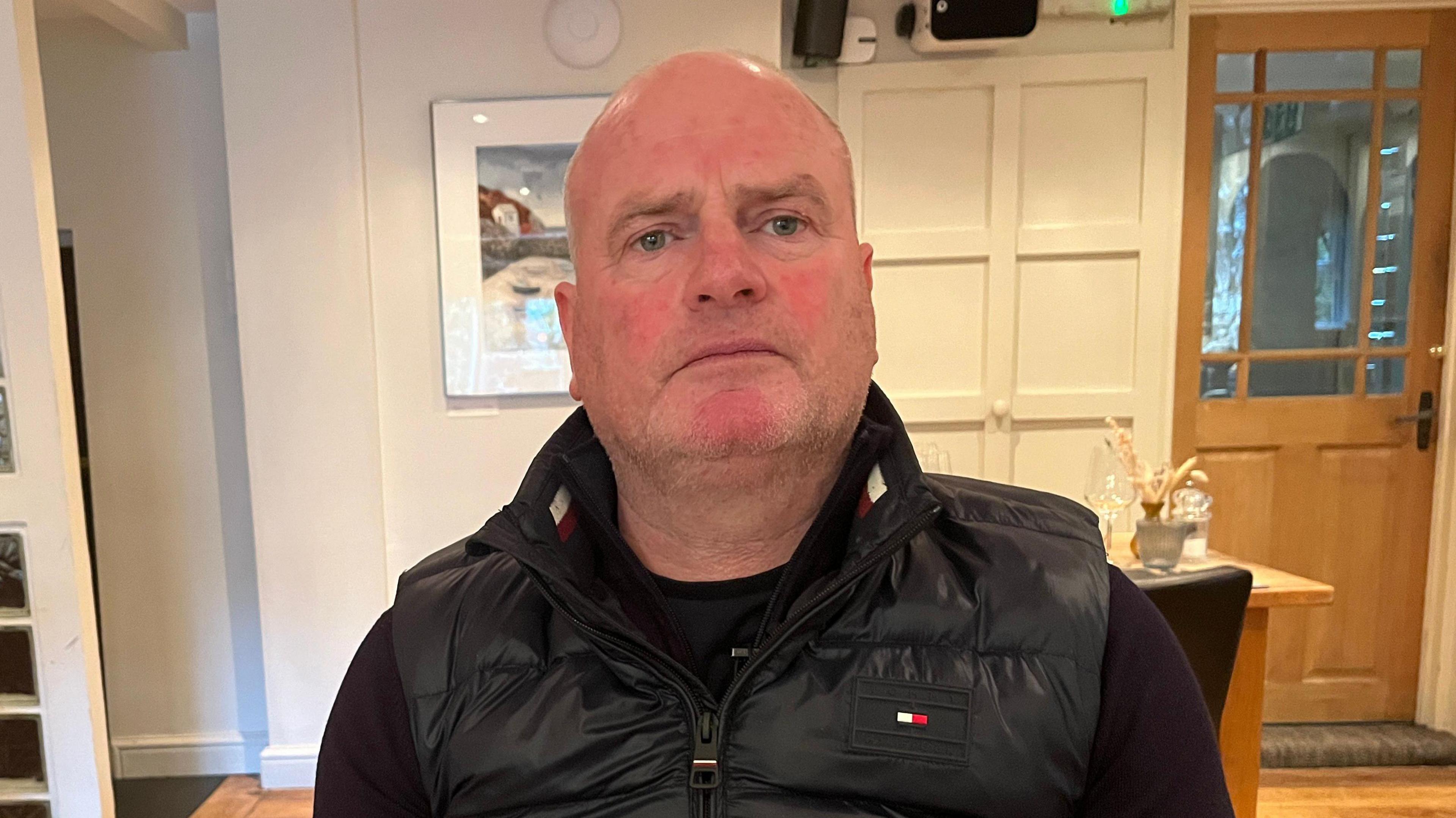 Llun o ddyn yn sefyll mewn tafarn yn edrych ar y camera. Mae'n gwisgo crys-t a gilet du. 