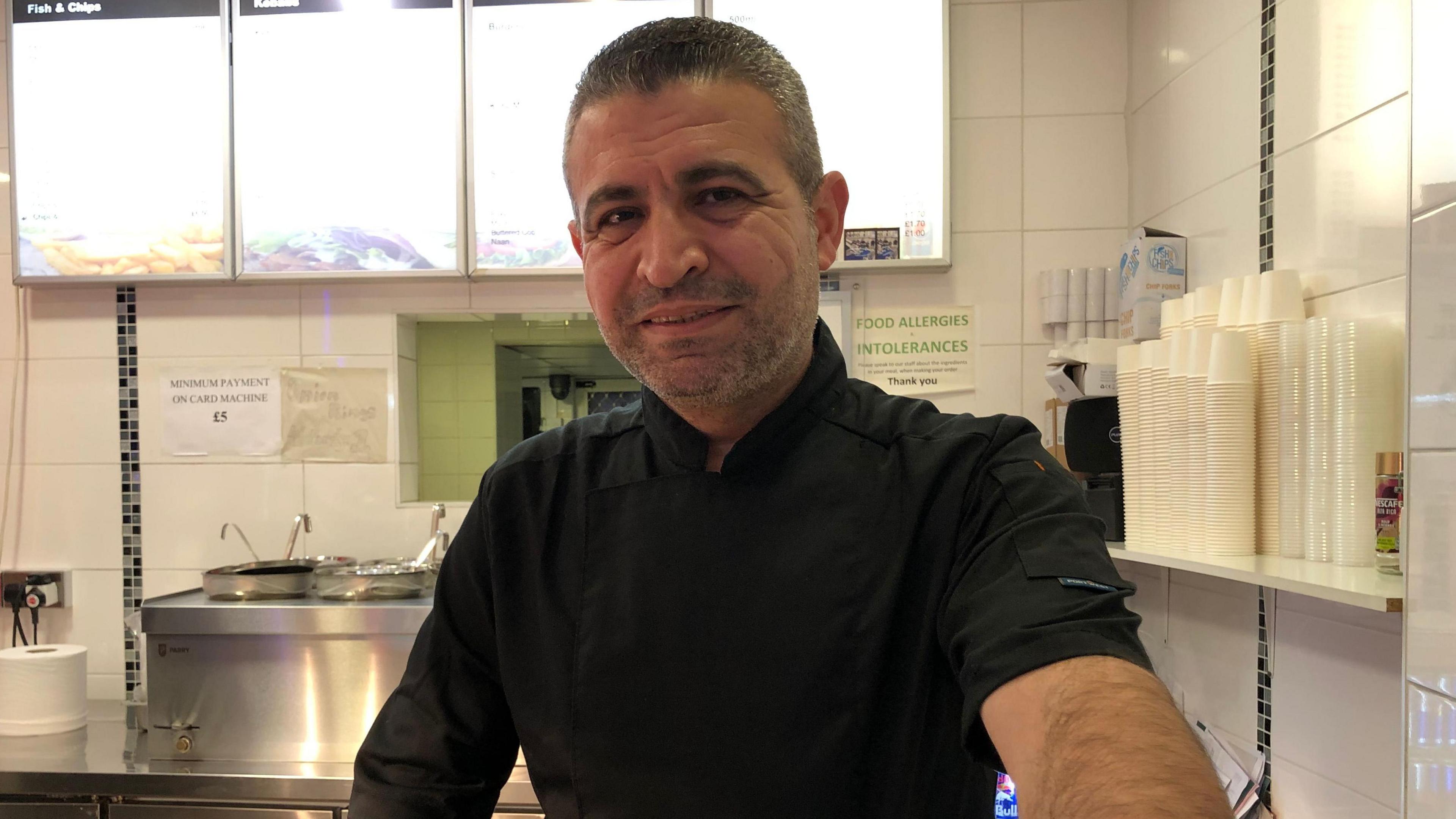 Sam Abbas, a worker at the Lace Market Chip Shop in Nottingham, standing at the counter in front of a menu and a shelve of paper takeaway containers. 