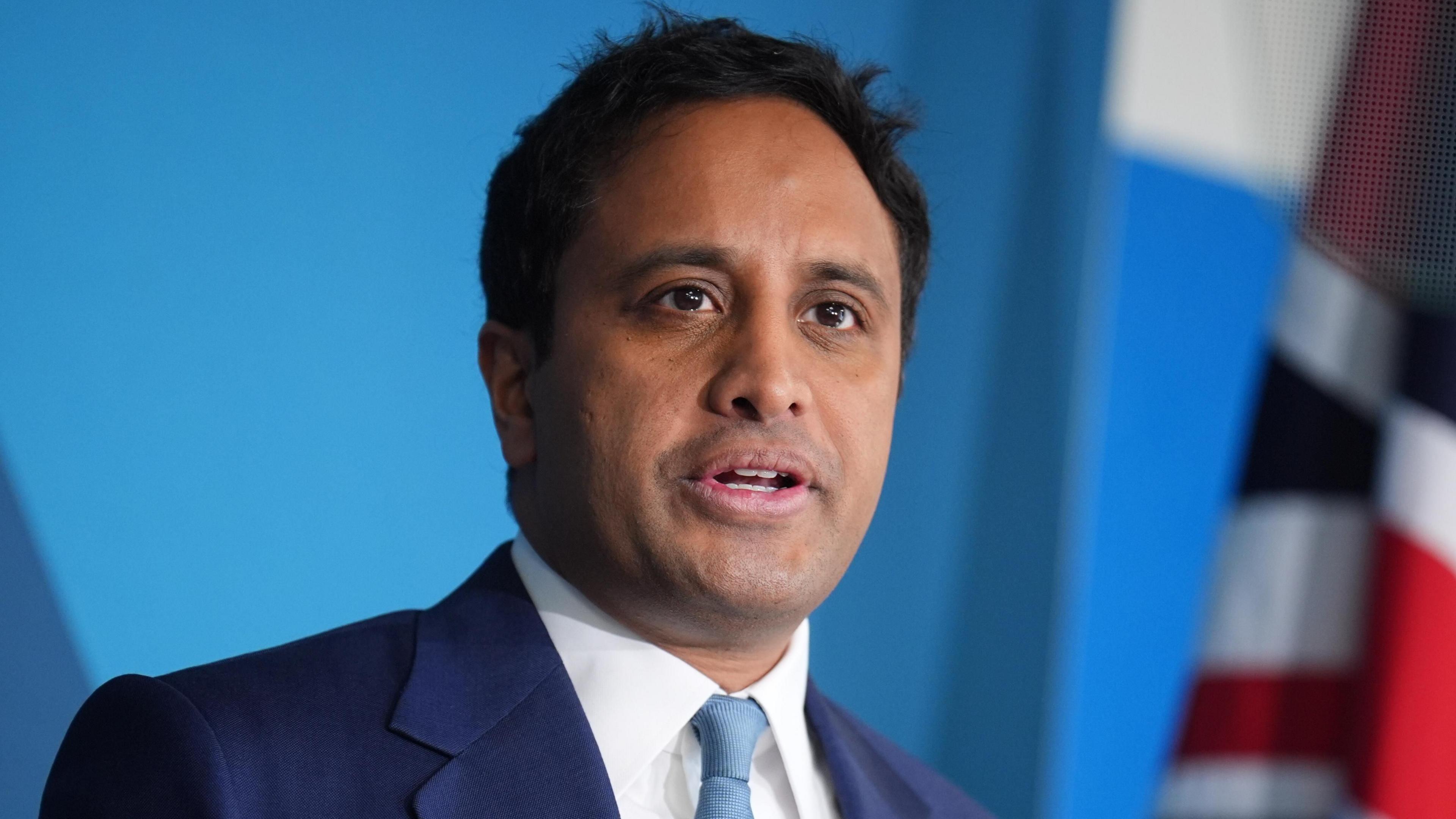 Zia Yusuf is stood in front of a blue background and a Union flag can be partially seen. He is wearing a dark blue suit, white shirt and a turquoise-coloured tie. He has short black hair.