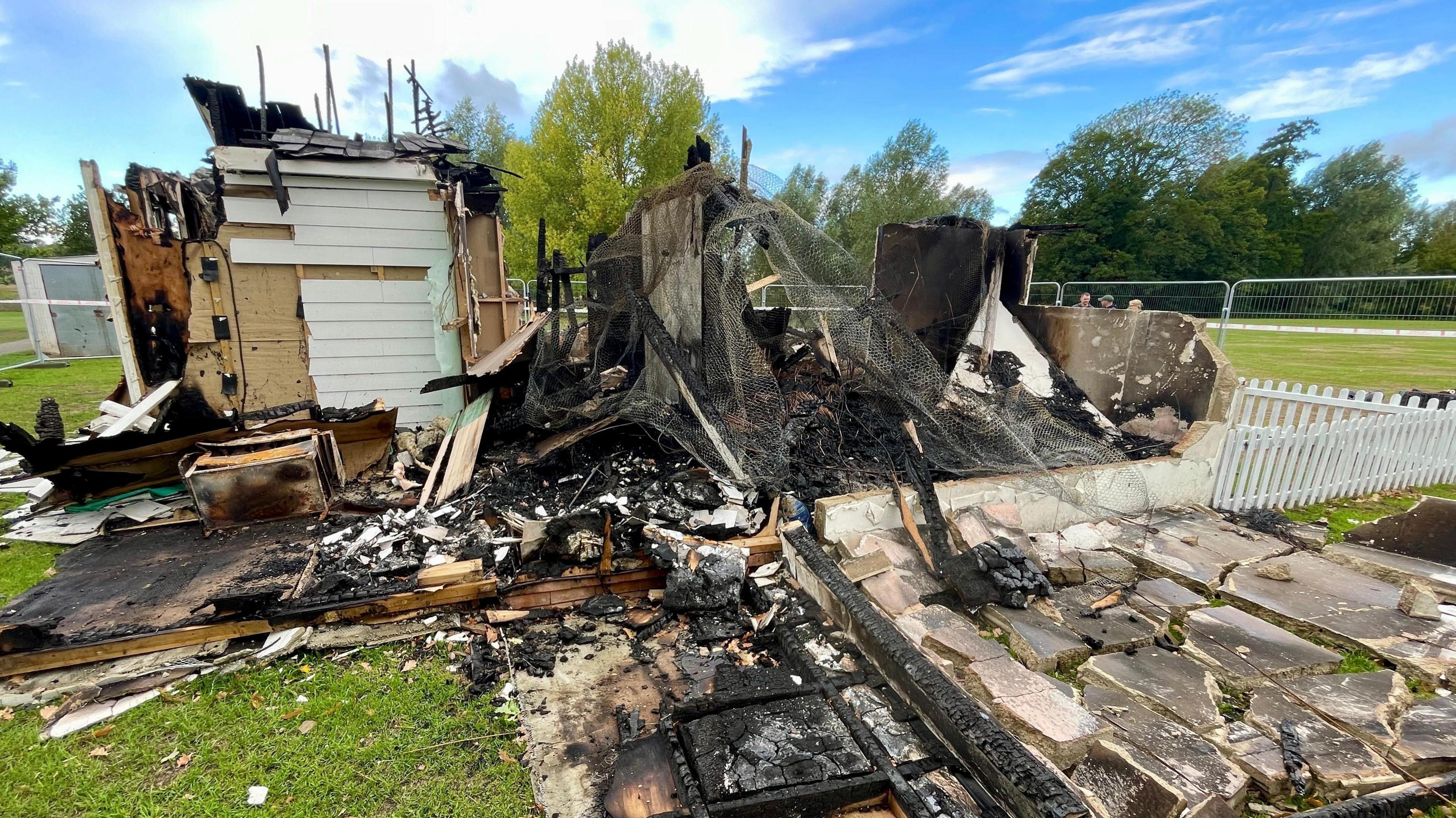 The remains of a white building on a grassy field. A fire has made half of the building collapse, including its roof, which has left ashes.