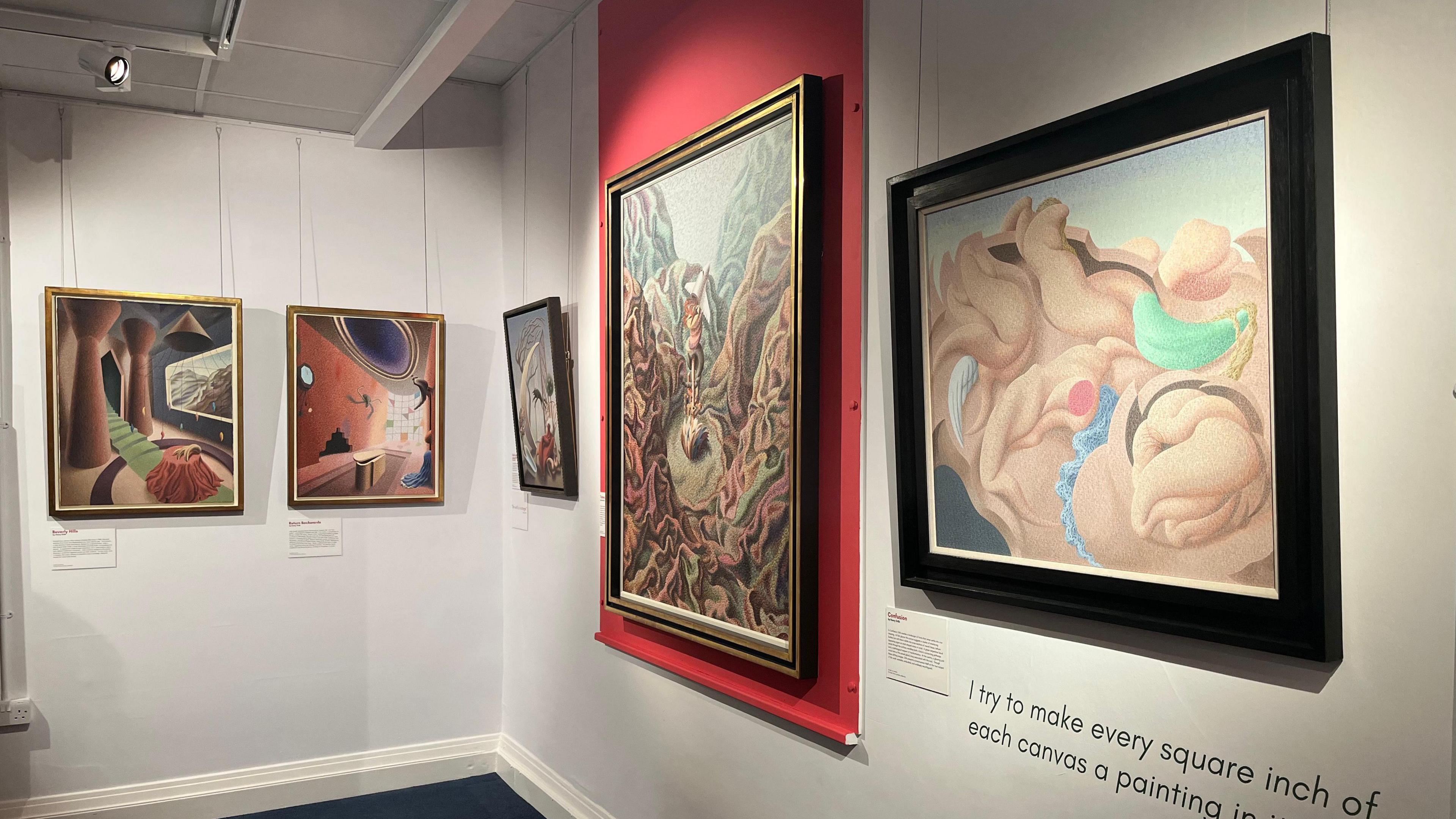 A corner of the gallery space, with five paintings in shot. All are surrealist with vibrant colours, unusual shapes and curves. All very neatly and precisely painted. backdrop is mostly a white wall with a bright red section behind the largest one