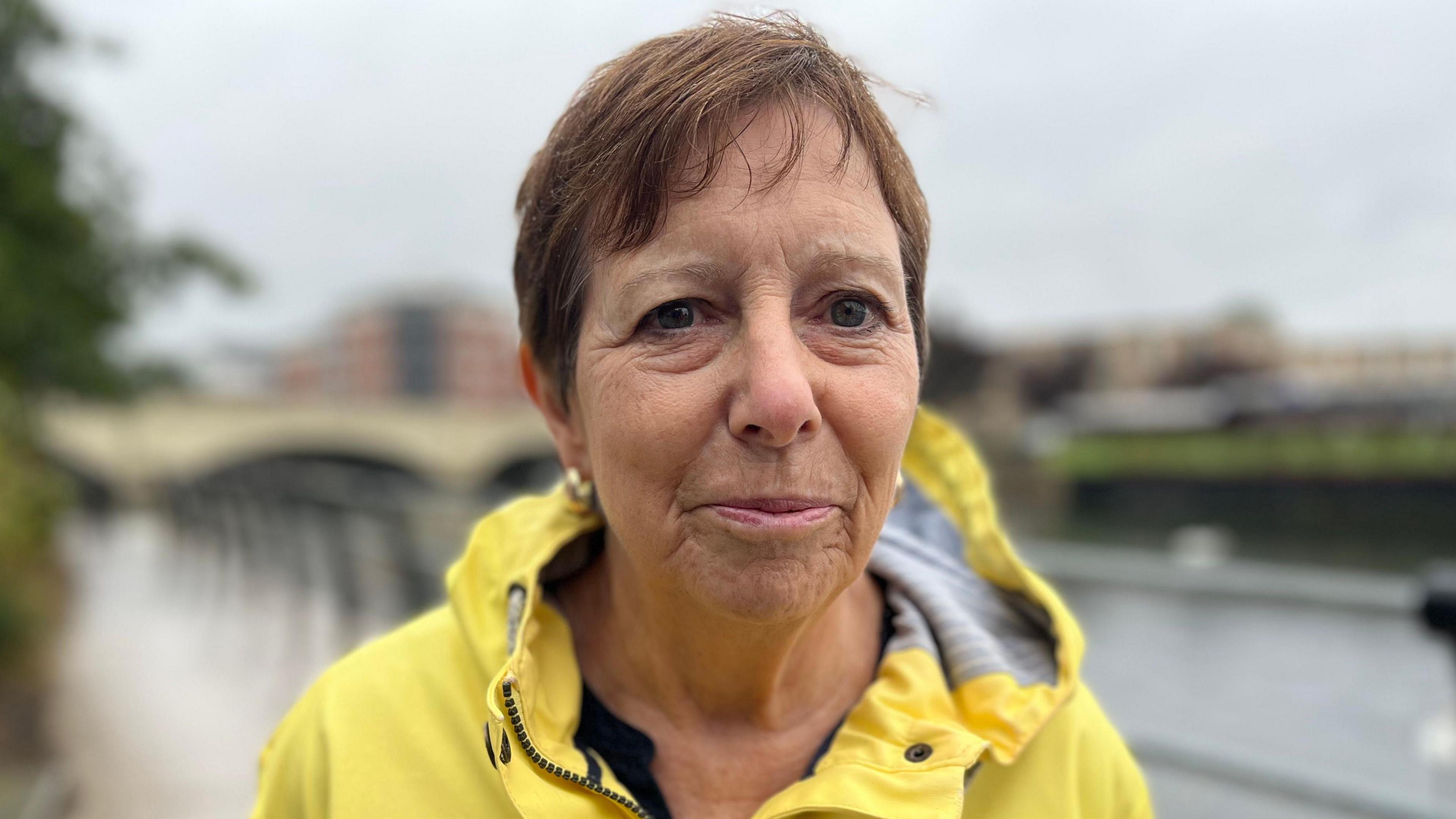 A lady with short brown hair is stood in the middle of the frame looking towards the camera with a light smile. She was wearing a yellow coat with a hoot and is stood in front of a small river.