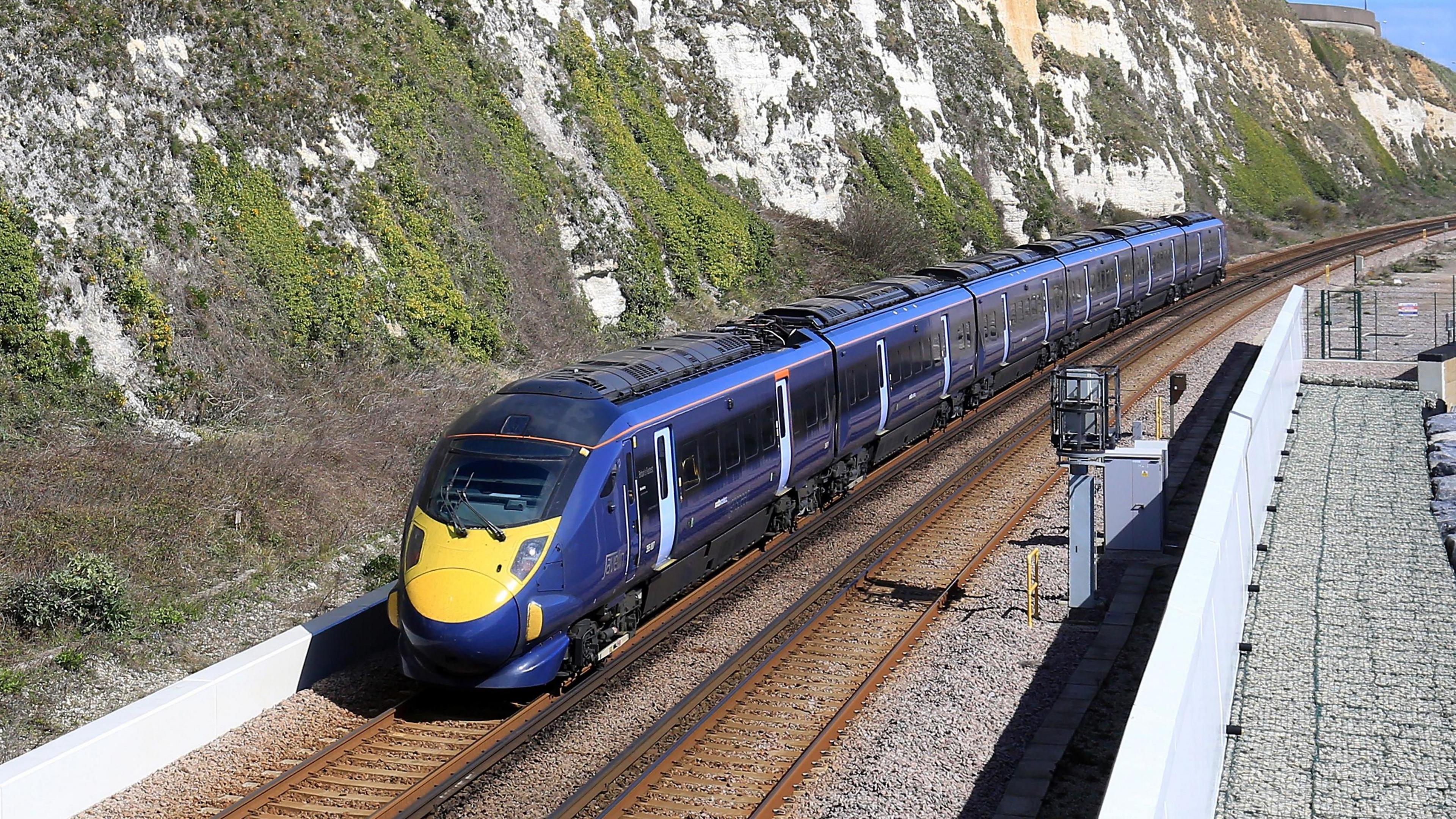 A purple and blue train running along tracks outside. The tracks are next to almost vertical cliff faces, which are white chalk, partially covered with some vegetation. The cliffs loom over the left of the image and stretch into the distance. On the right of the image is some rail infrastructure, what appears to be some hardcore and signalling equipment. It is a bright day and there is a small amount of blue sky visible in the top right hand corner of the image.