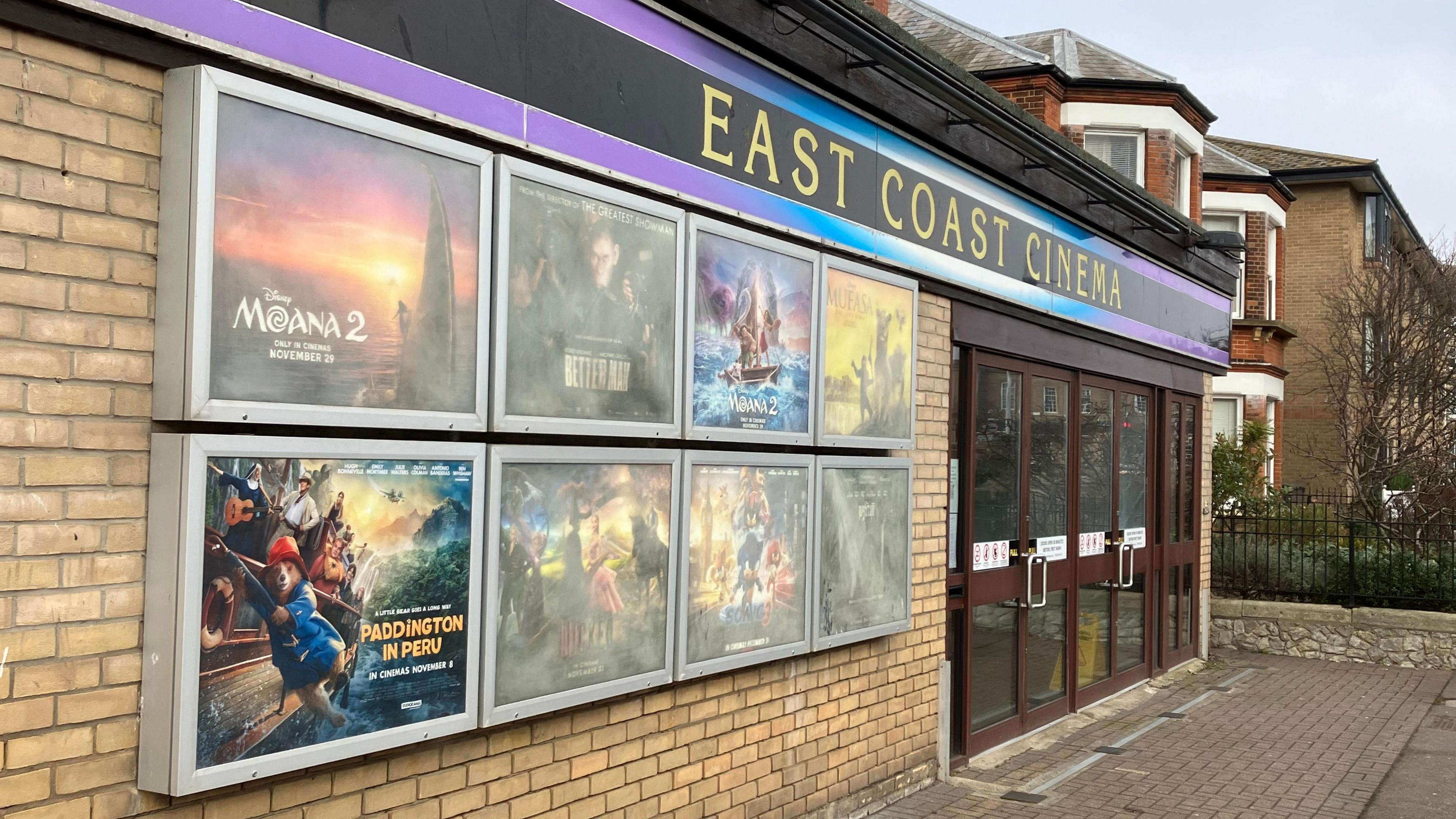 The facade of East Coast Cinema with pale bricks, brown doors and eight metal and glass cases containing a variety of film posters