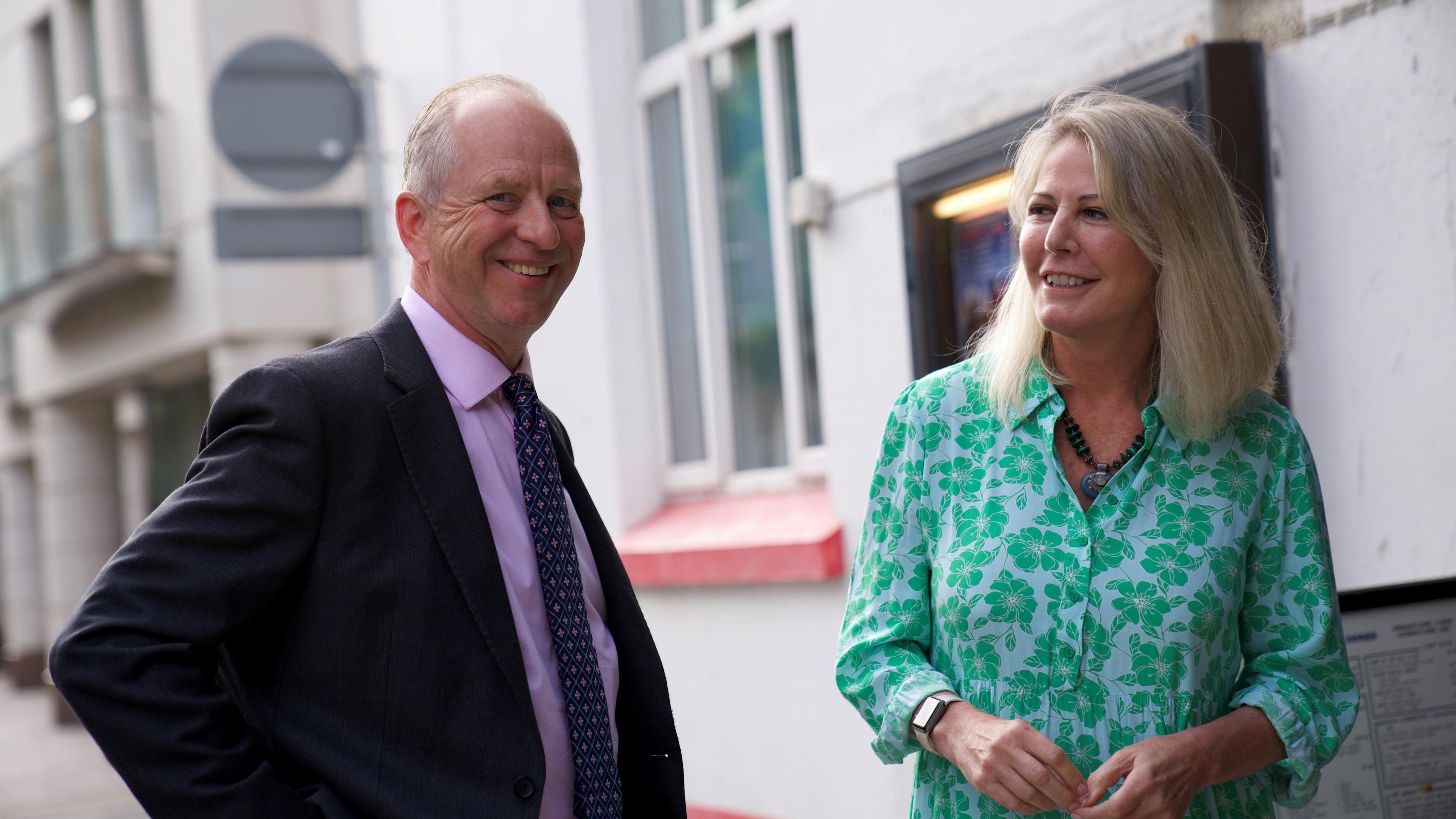 Vice Admiral Jerry Kyd CBE pictured smiling with Jersey’s Minister for International Development, Deputy Carolyn Labey. Deputy Labey is wearing a green and white shirt, she has blonde hair. Vice Admiral Kyd is wearing a pink suit shirt, a navy blue patterned tie and a black blazer on top.