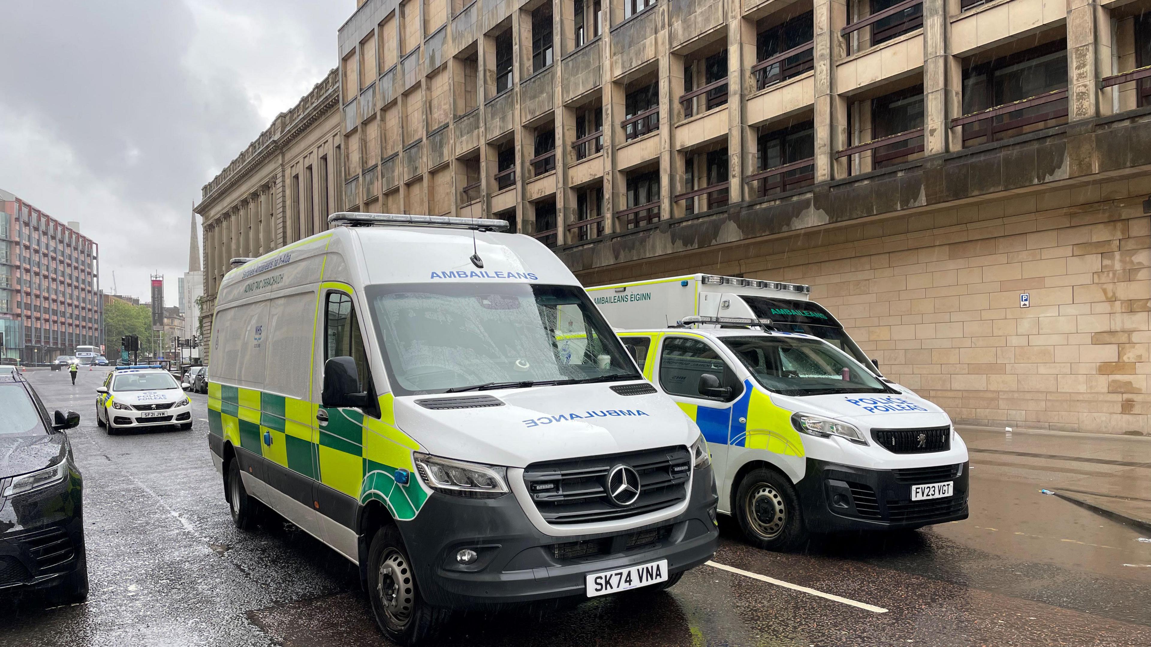 A police van, car and ambulance sitting outside the Mitchell library building, with a police officer walking around the scene in the distance. 