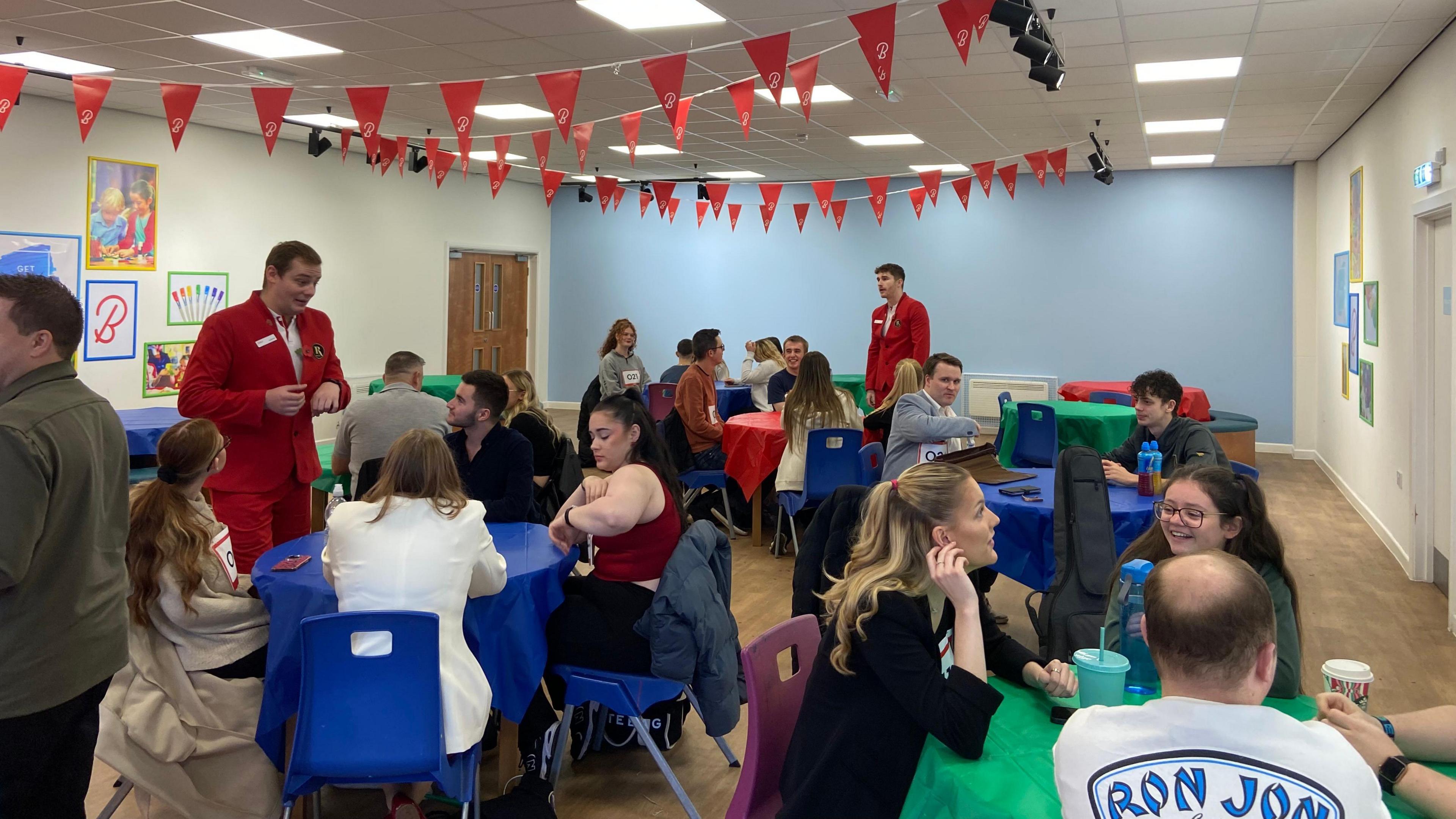 People wearing numbers on their chests sit around tables chatting ahead of auditions. Two mean dressed in red suits walk among them. Red and black bunting hangs from the ceiling.