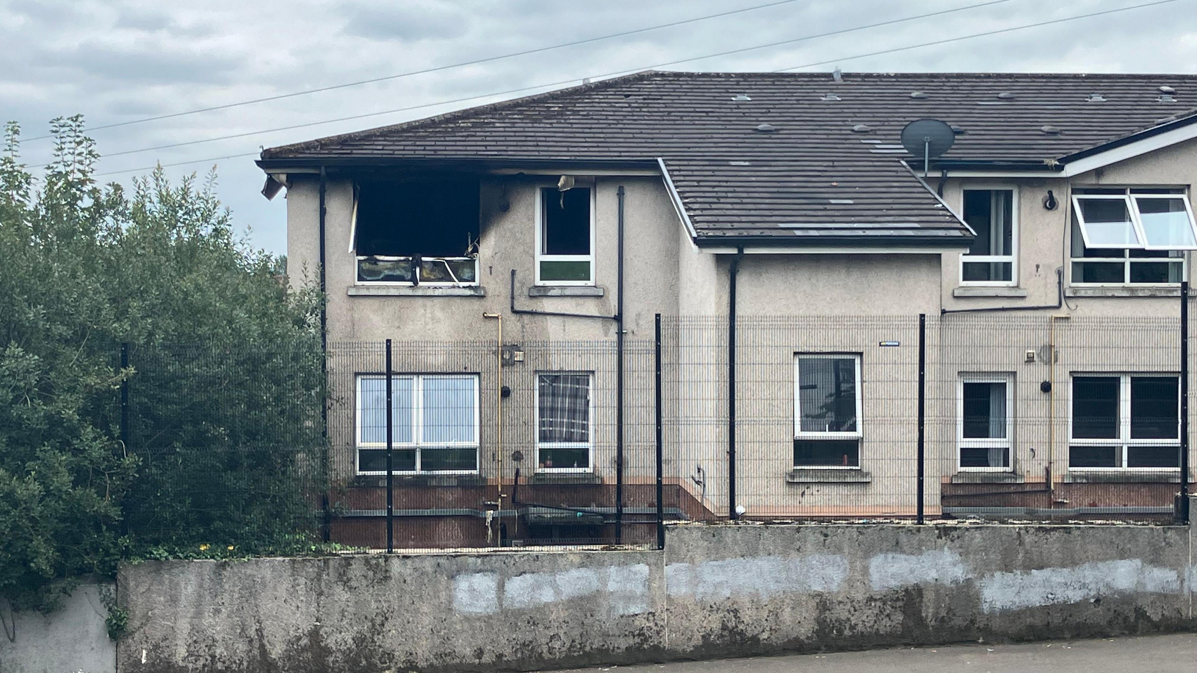 A cream coloured block of flats with a brown tiled roof and white windows. One of the flats has been set alight. Its' windows are smashed and there is smoke damage. There is a stone wall with black metal railing around the property as well, and trees along the side.