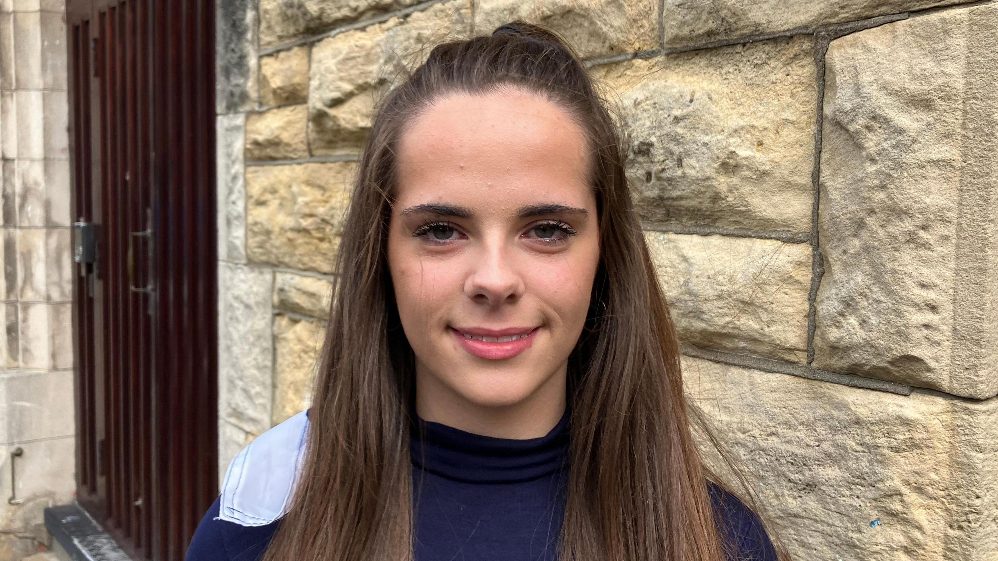Erin has long brown hair. She is smiling with hair tied back.
She is wearing a navy dancer outfit and is standing beside Derry's historic walls.