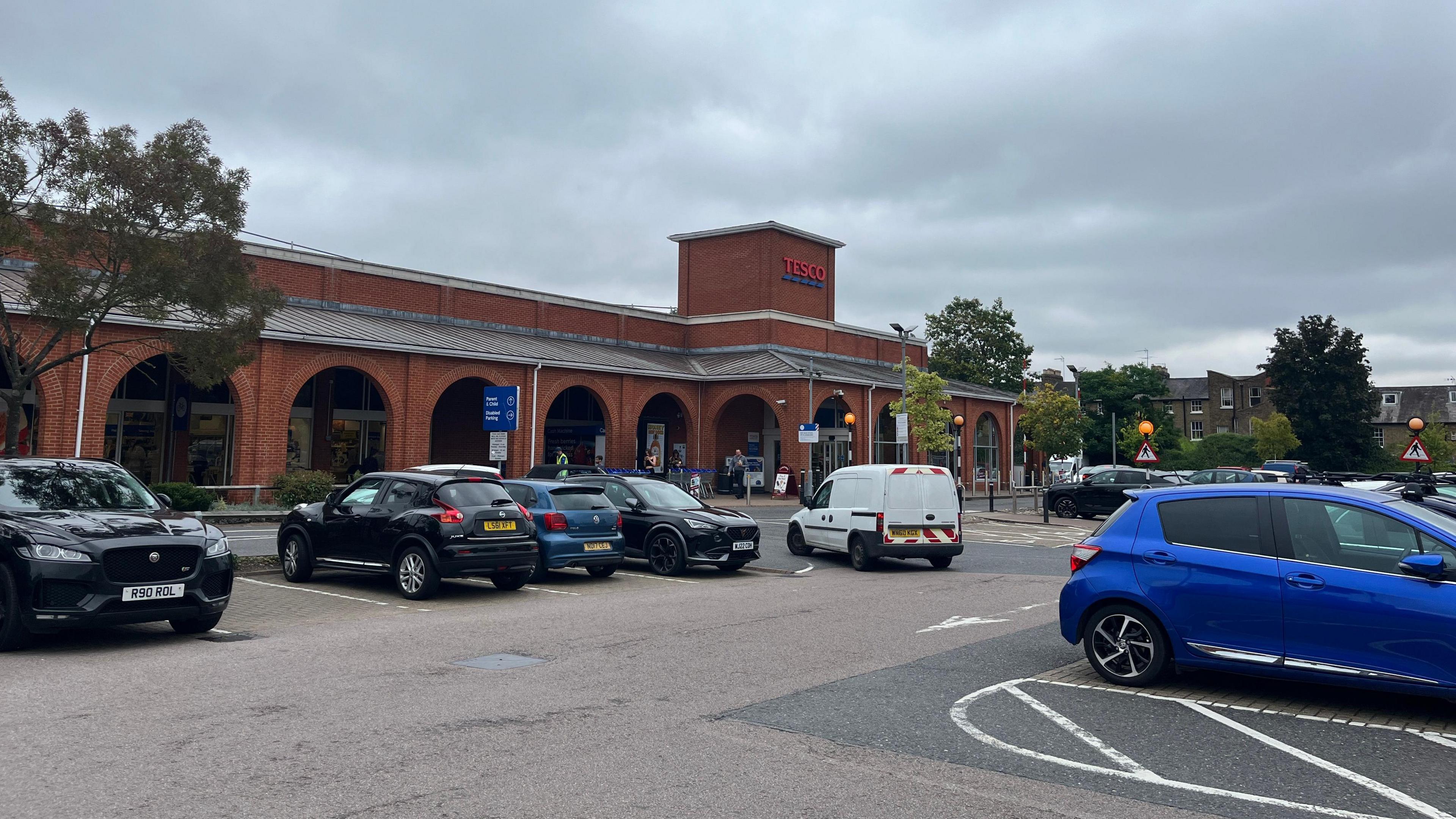 A car park with mostly blue and black cars outside a red-brick Tesco supermarket