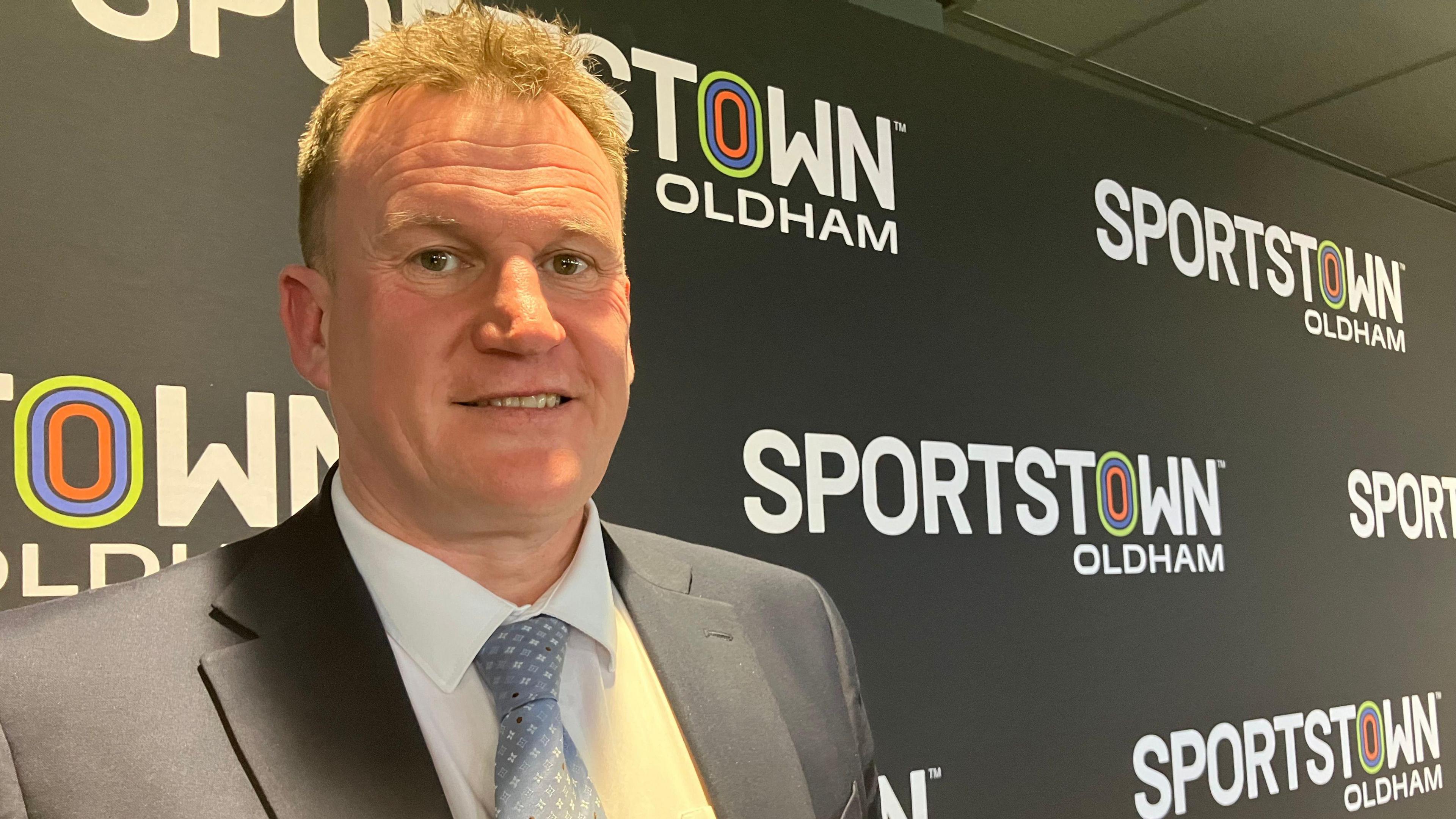 Darren Royle, who is wearing a suit, smiles next to a black hoarding bearing the branding 'Sportstown Oldham' in white lettering. 