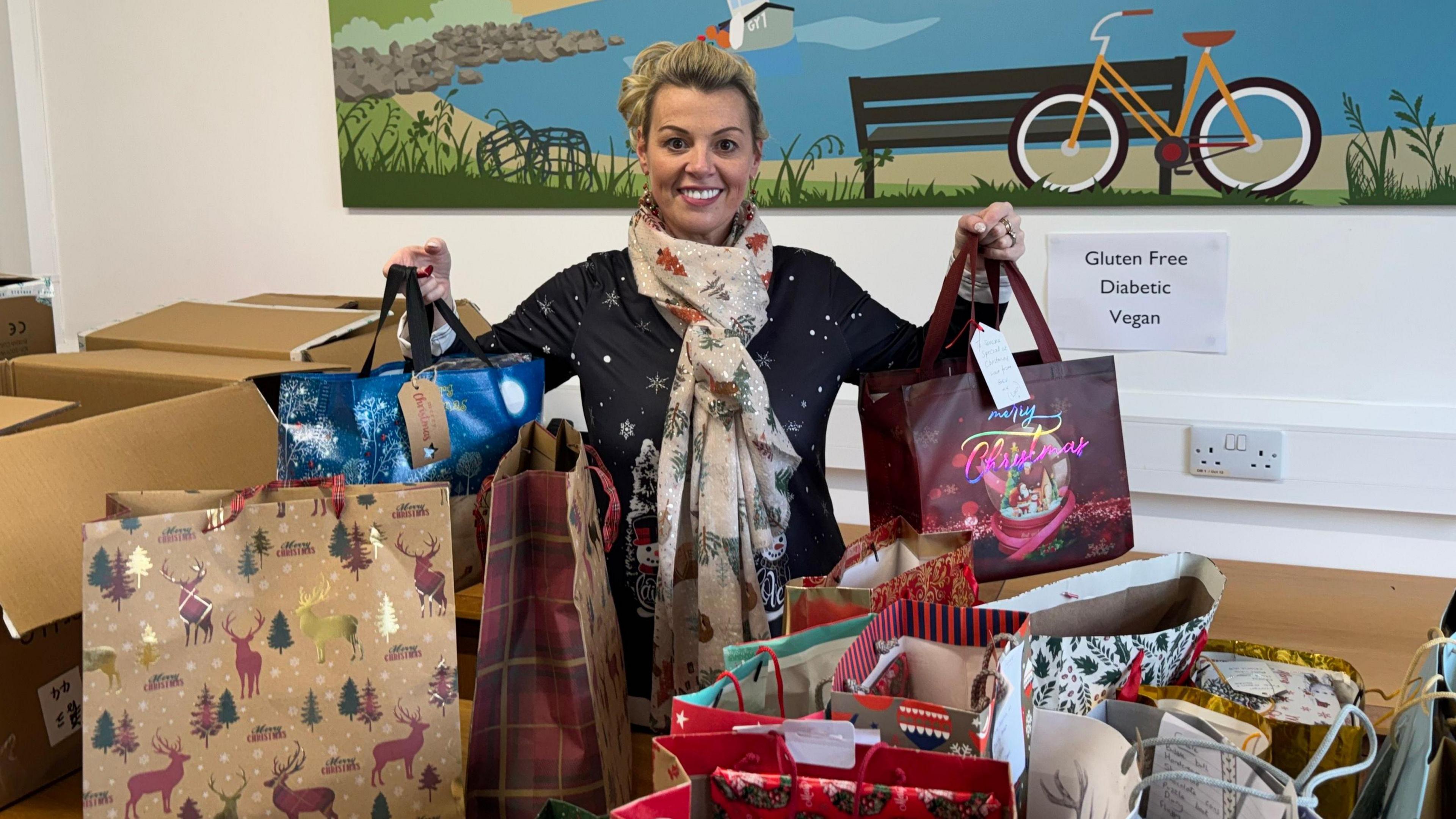 Donna Patch, chairman of gifts for elderly and vulnerable charity, holding a gift bag in each hand. Ms Patch is stood in front of a pile of other gift bags. She has light hair and is wearing a navy Christmas top. She has a Christmas scarf around her neck. Ms Patch has her hair tied up and is smiling at the camera.