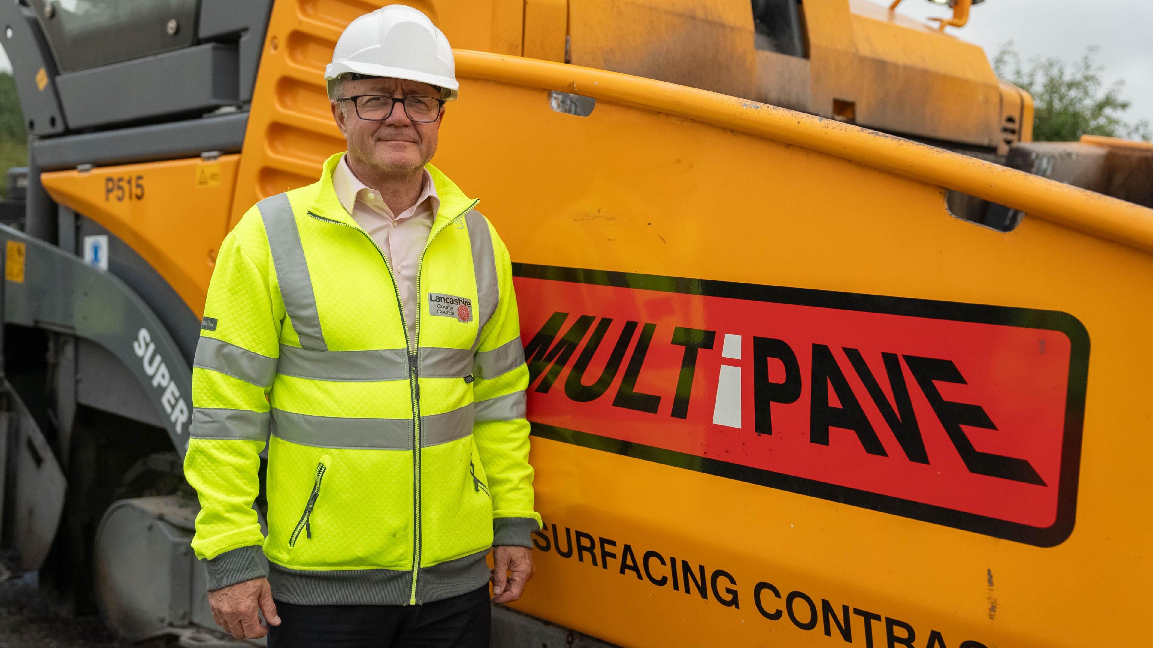 Councillor Rupert Swarbrick is smiling as she stands next to a roadworks vehicle. He is wearing a hi-vis jacket with the Lancashire County Council logo on it, along with a safety helmet.