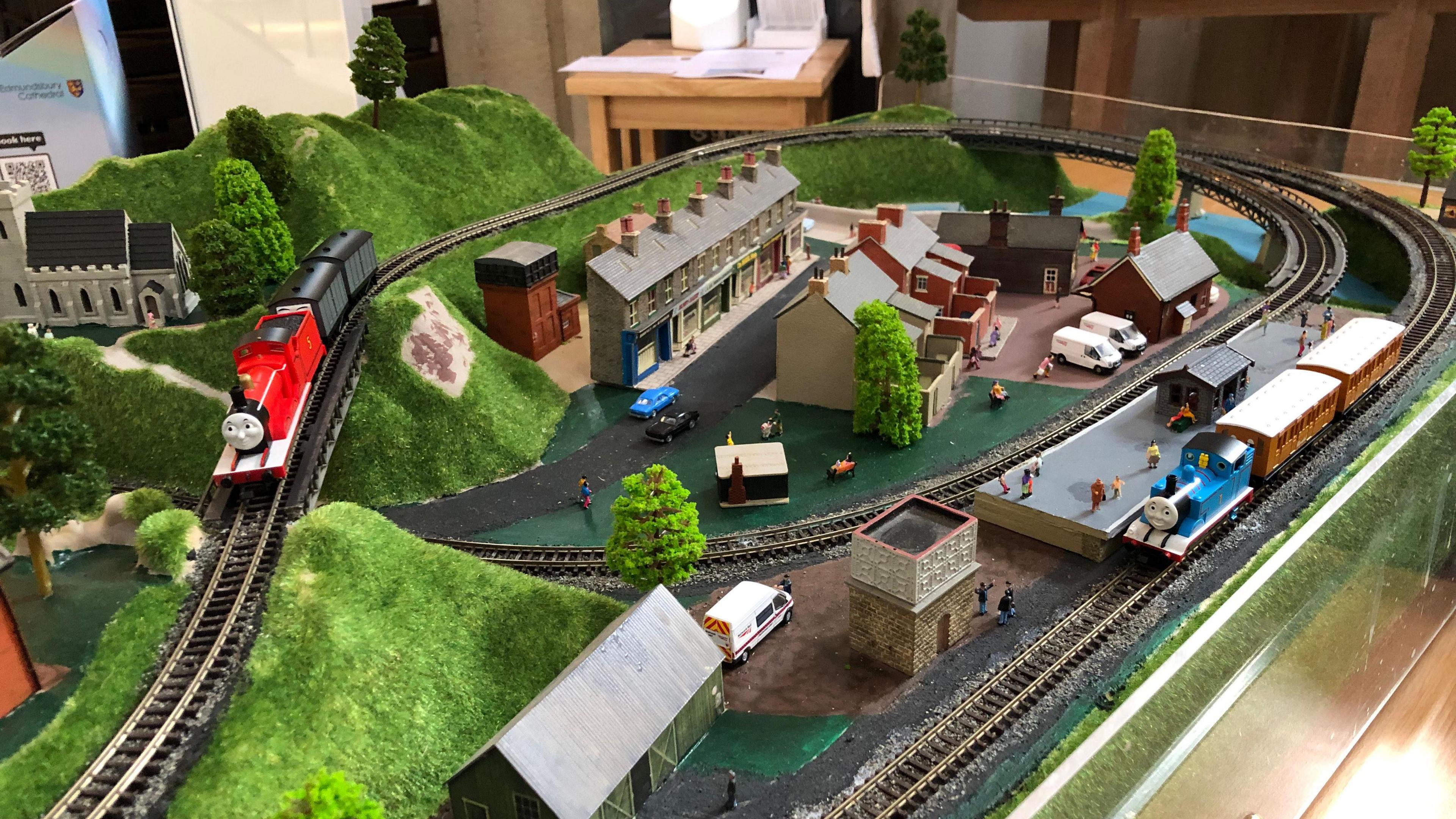 A close up picture of a Thomas the Tank Engine themed model railway track. Railway tracks can be seen with model trains from the show on top of them. Model buildings lie around the track as well as road vehicles and small models of people. 