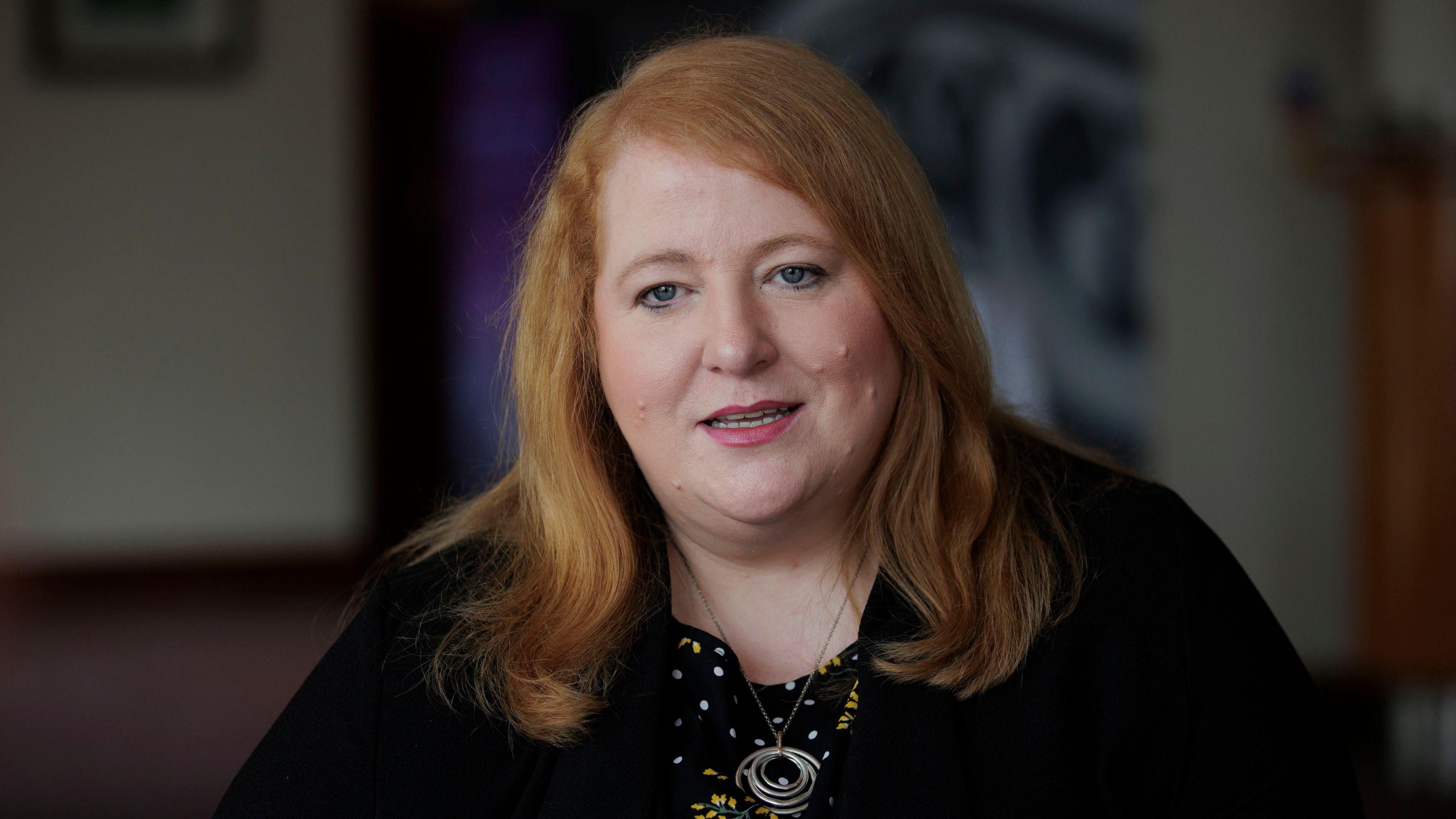 Naomi Long looking at the camera. She is wearing a black top and a silver necklace. The background is out of focus.