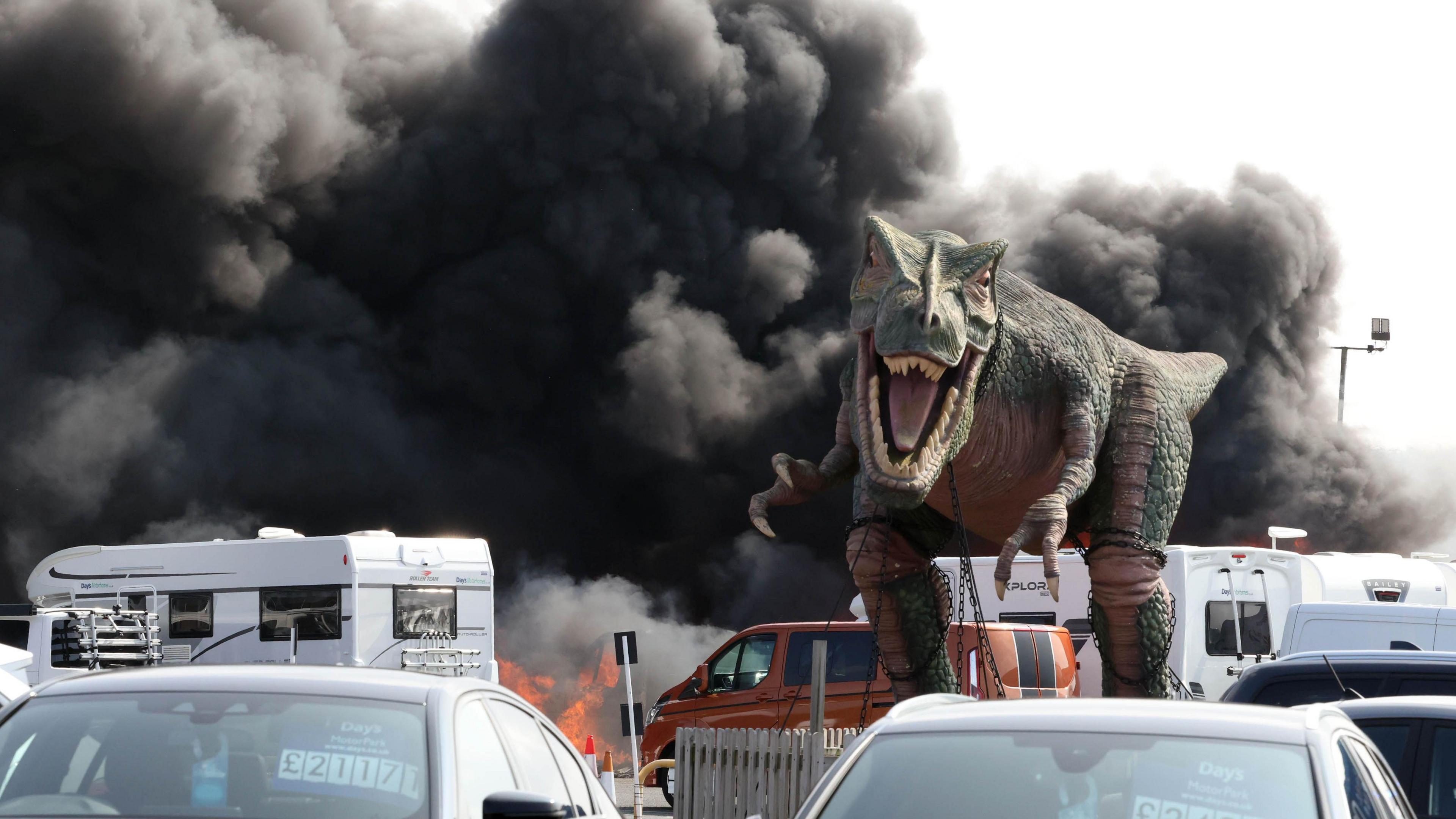 A dinosaur model stood in frozen roar on a car forecourt as smoke rose into the sky behind it