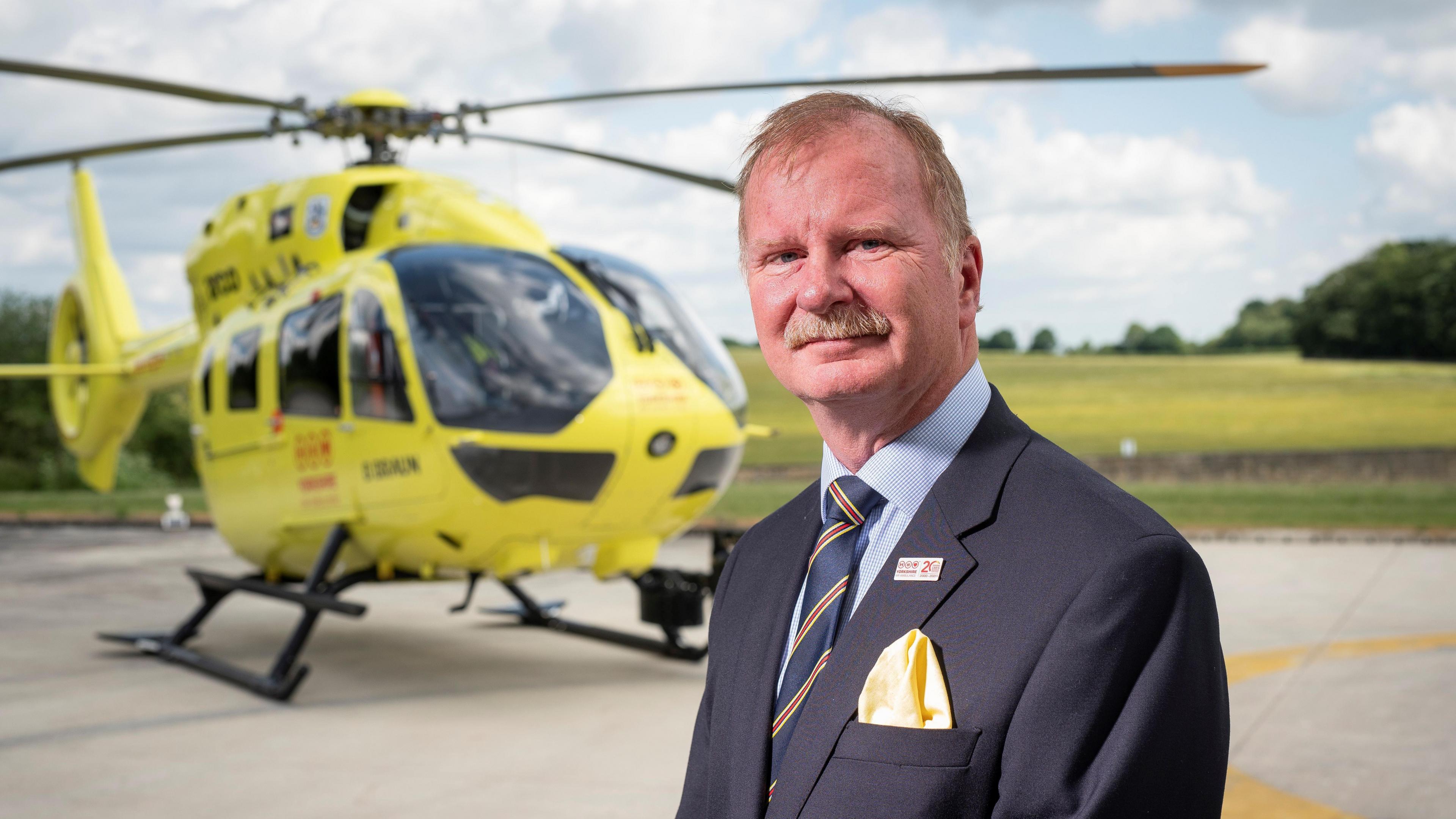 Mike Harrop standing in front of yellow helicopter.
Mike is wearing a blazer with a yellow pocket square and a navy blue tie with red and yellow stripes. He is wearing a light blue and white checked shirt and a blazer with a tag on.