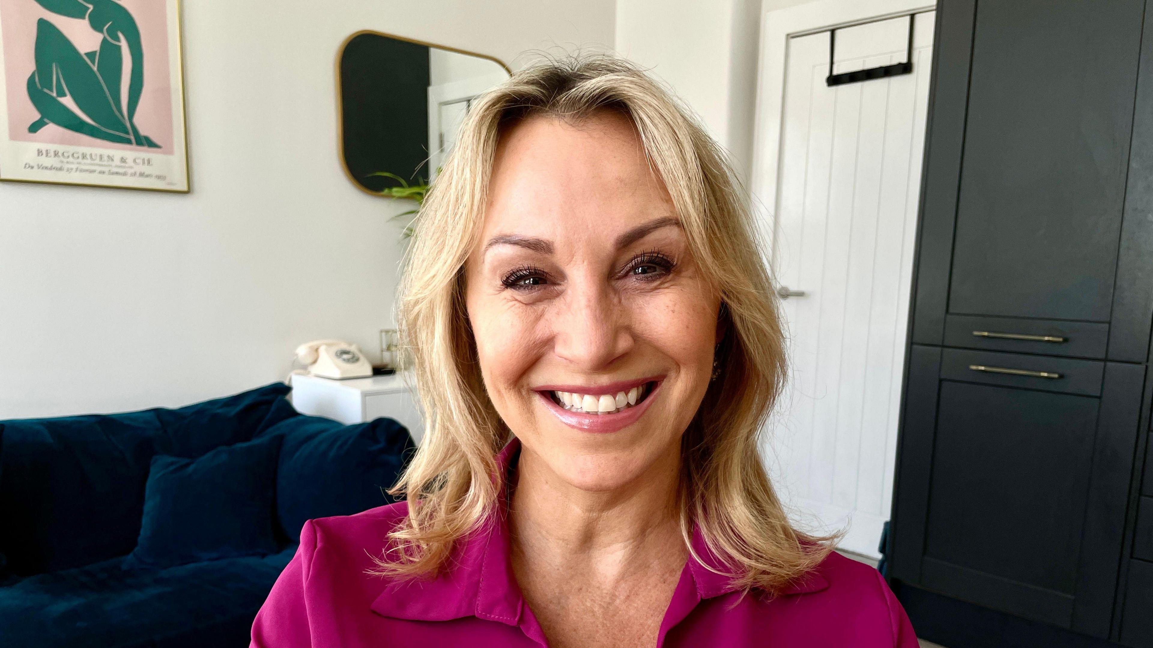 A woman with blonde hair and pale pink lipstick smiles widely at the camera