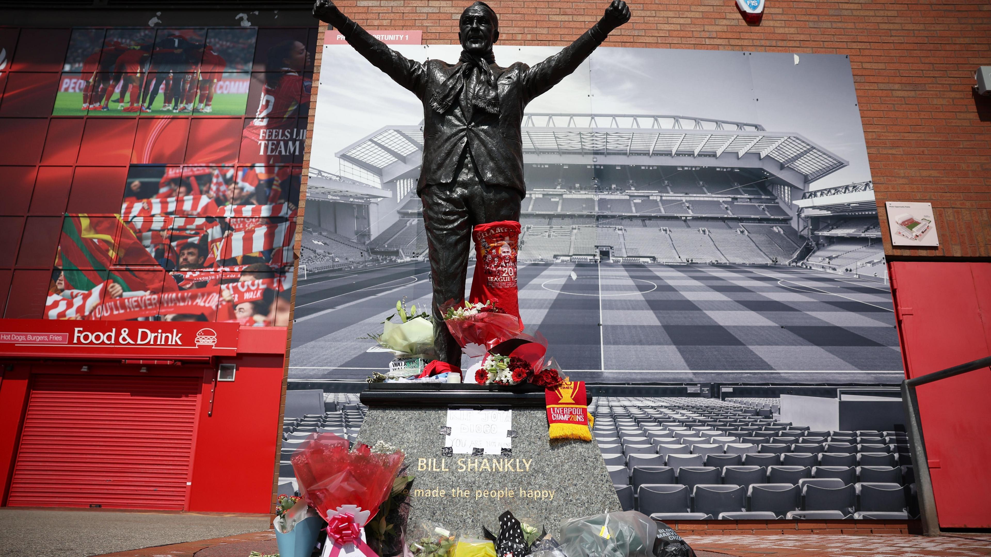 A statue of Bill Shankly on a stone podium is surrounded by flowers and scarves, with the stadium in the backdrop